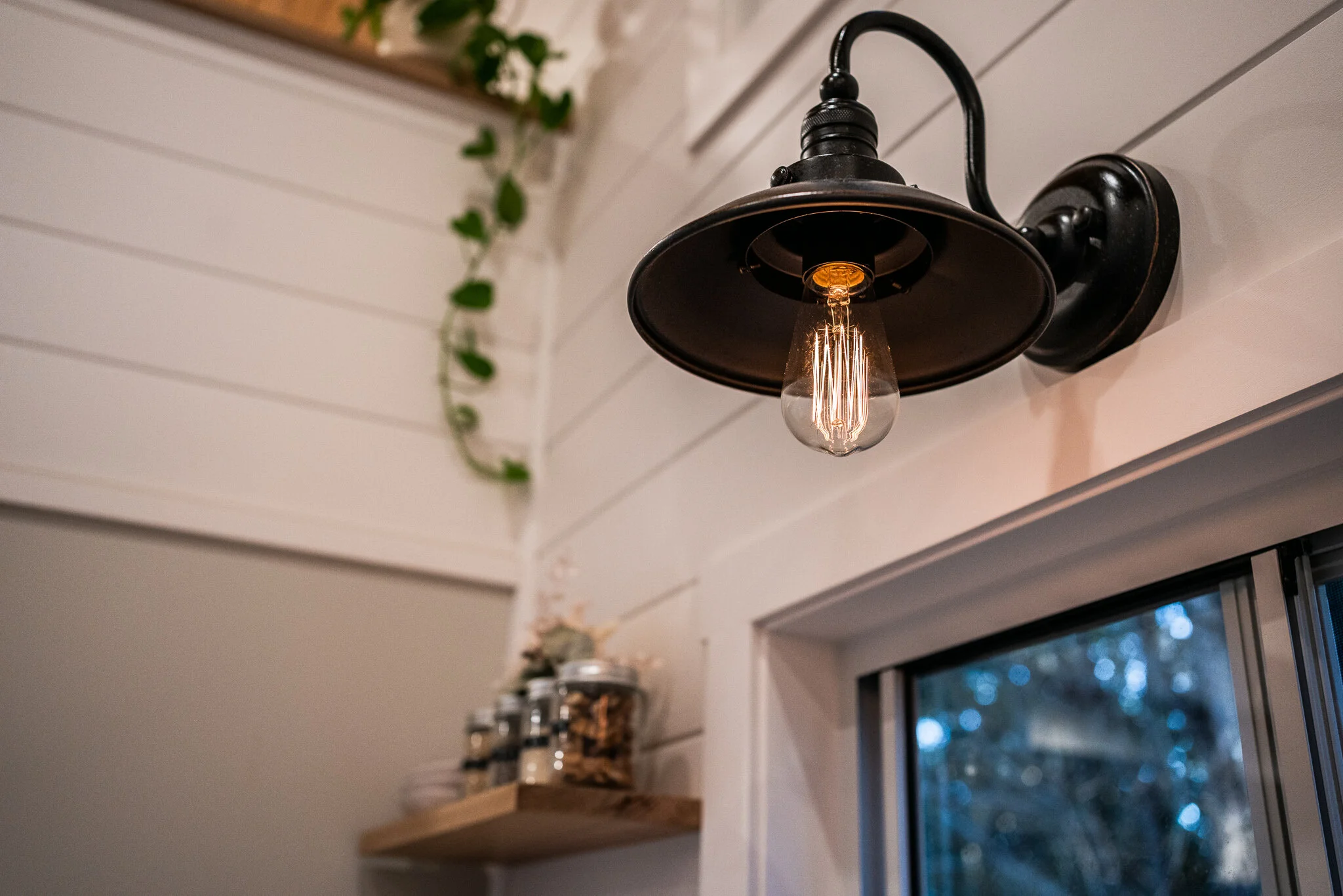 Wall-mounted black metal sconce with an Edison bulb, next to a window. Nearby, a wooden shelf holds glass jars and a small potted plant; a vine hangs above. The walls are white with horizontal panels.