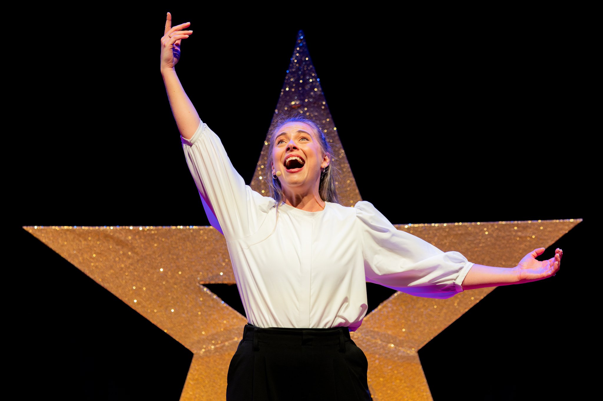 A woman on stage, smiling and singing, with a large gold glittery star behind her.
