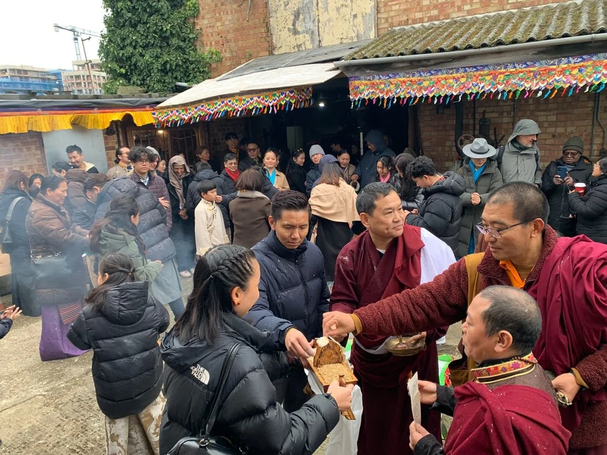 A gathering of people at an outdoor event with traditional Buddhist monks in maroon robes. Monks are giving out blessings or offerings, and everyone is dressed warmly, indicating cool weather. Colorful decorations hang from the covered area, and many people are taking photos or observing.