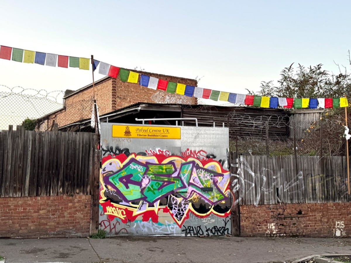 Graffiti art on a metal gate with a yellow sign above it reading "Polyul Centre UK Tibetan Buddhist Centre," surrounded by a wooden fence, barbed wire, and colorful prayer flags strung across the top of the fence.