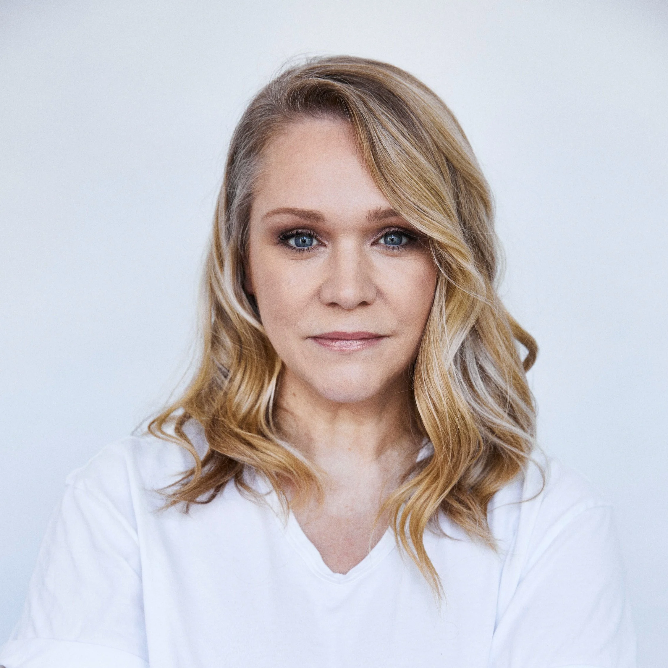 Ganzseitiges Porträt einer Frau mit blonden, gewellten Haaren, blauen Augen und hellem Make-up vor weißem Hintergrund.