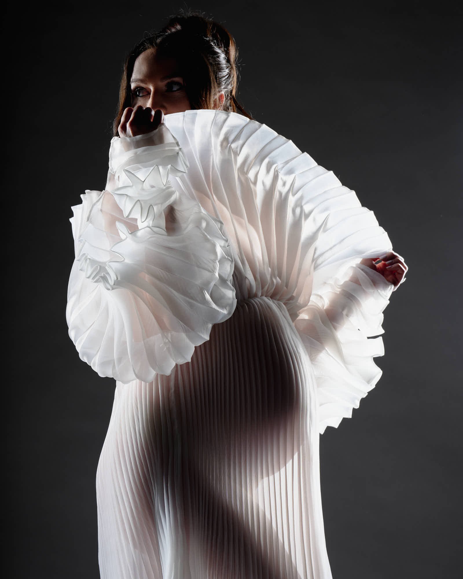 Sculptural white pleated gown with oversized ruffled sleeves against a dark studio backdrop.