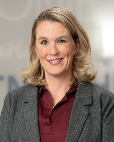 Best Midwifery winner portrait in a dark blazer, smiling, soft grey background.