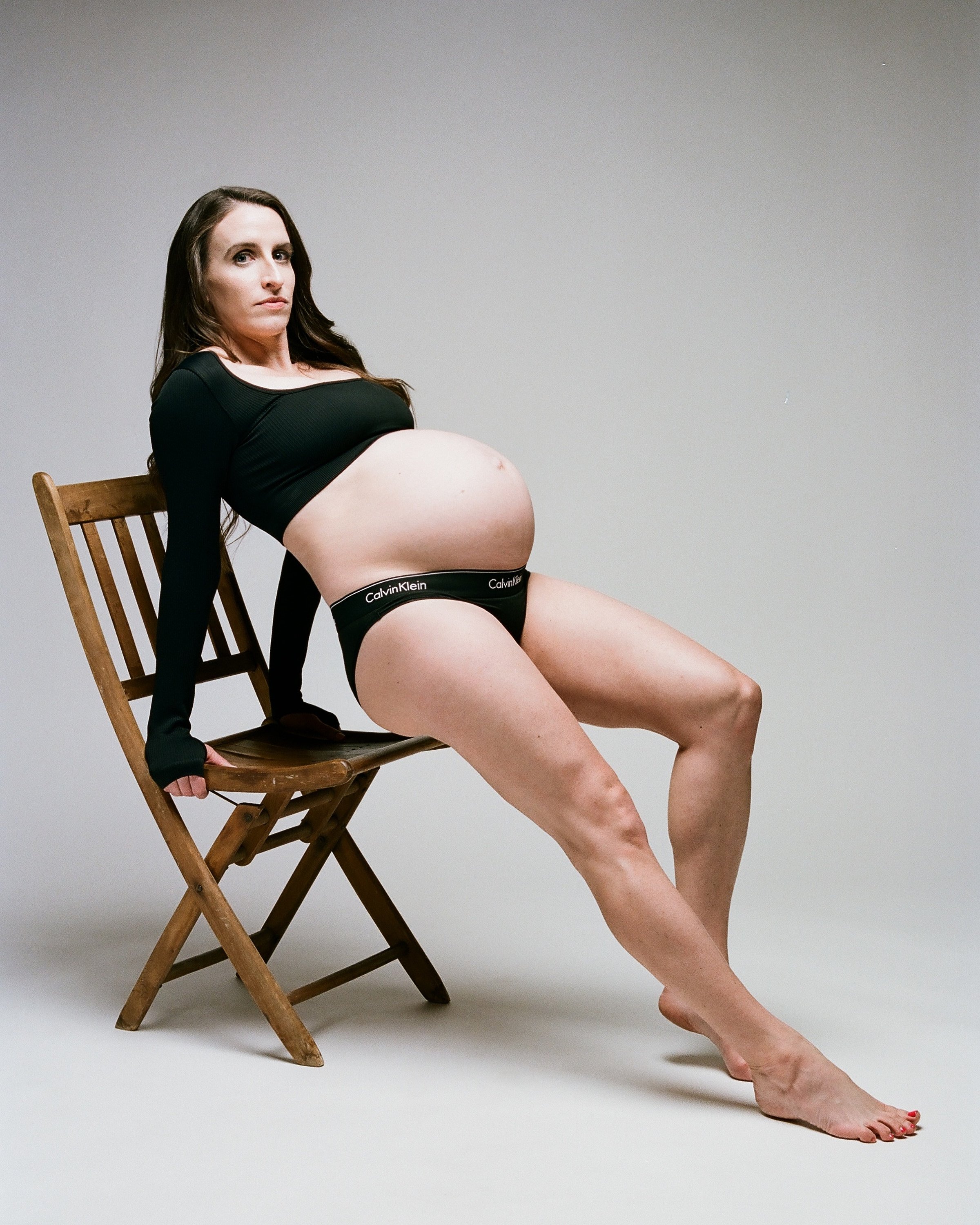 Seated back on a wooden chair with legs stretched long, black top pulled above the bare bump.