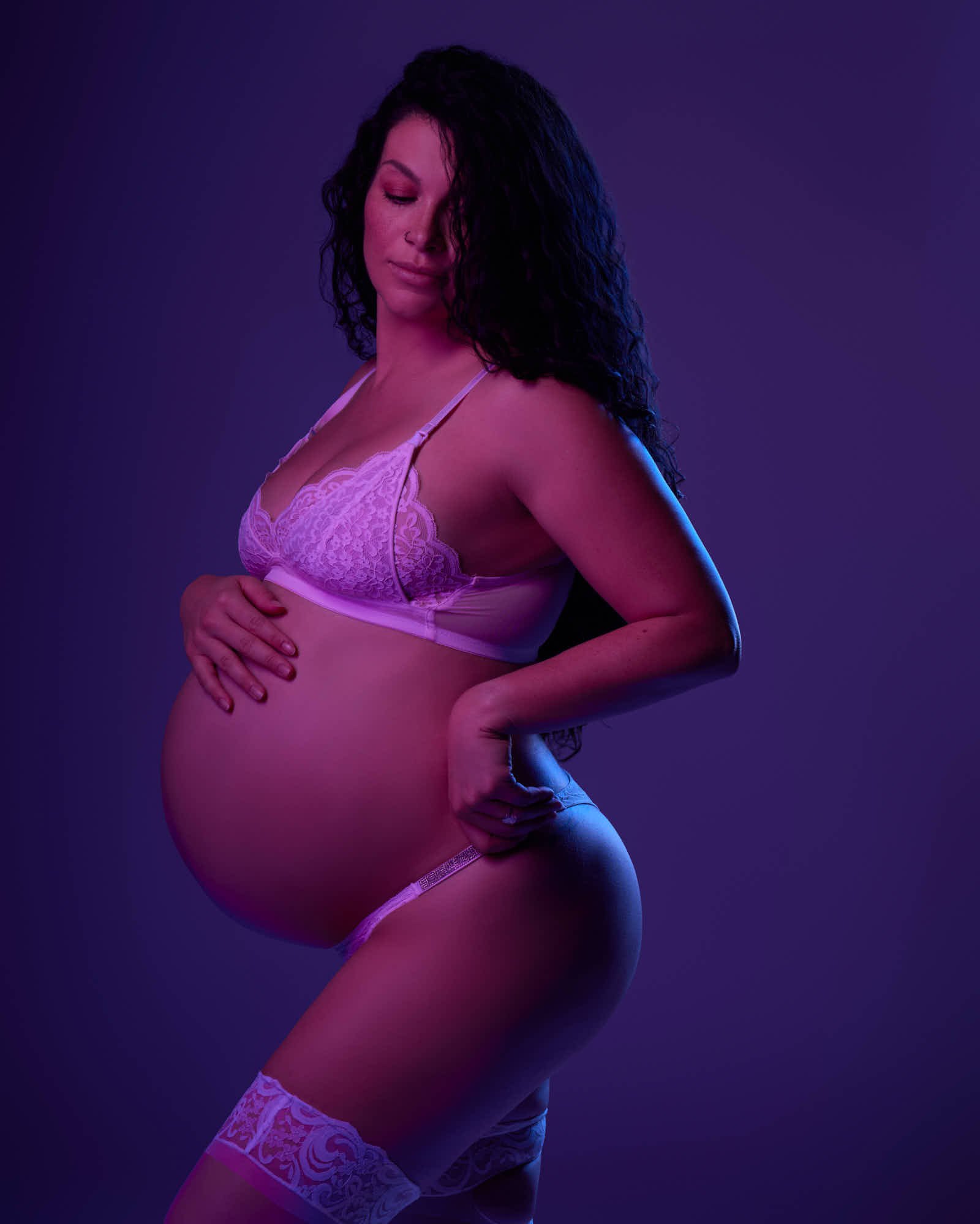 Purple lace bralette and thigh-high stockings under violet gel light, hands framing the bump.