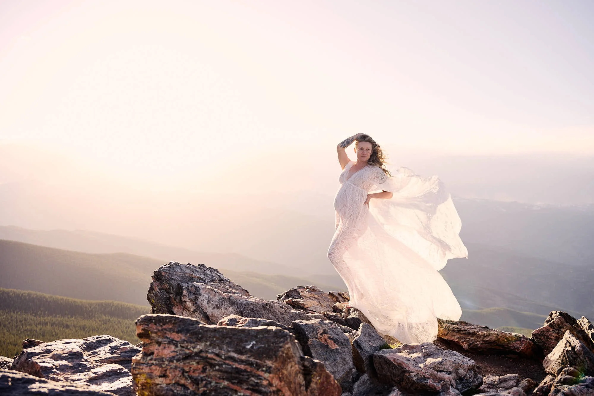 Arm raised in triumph on granite summit rocks, lace gown fanning above the hazy valley.