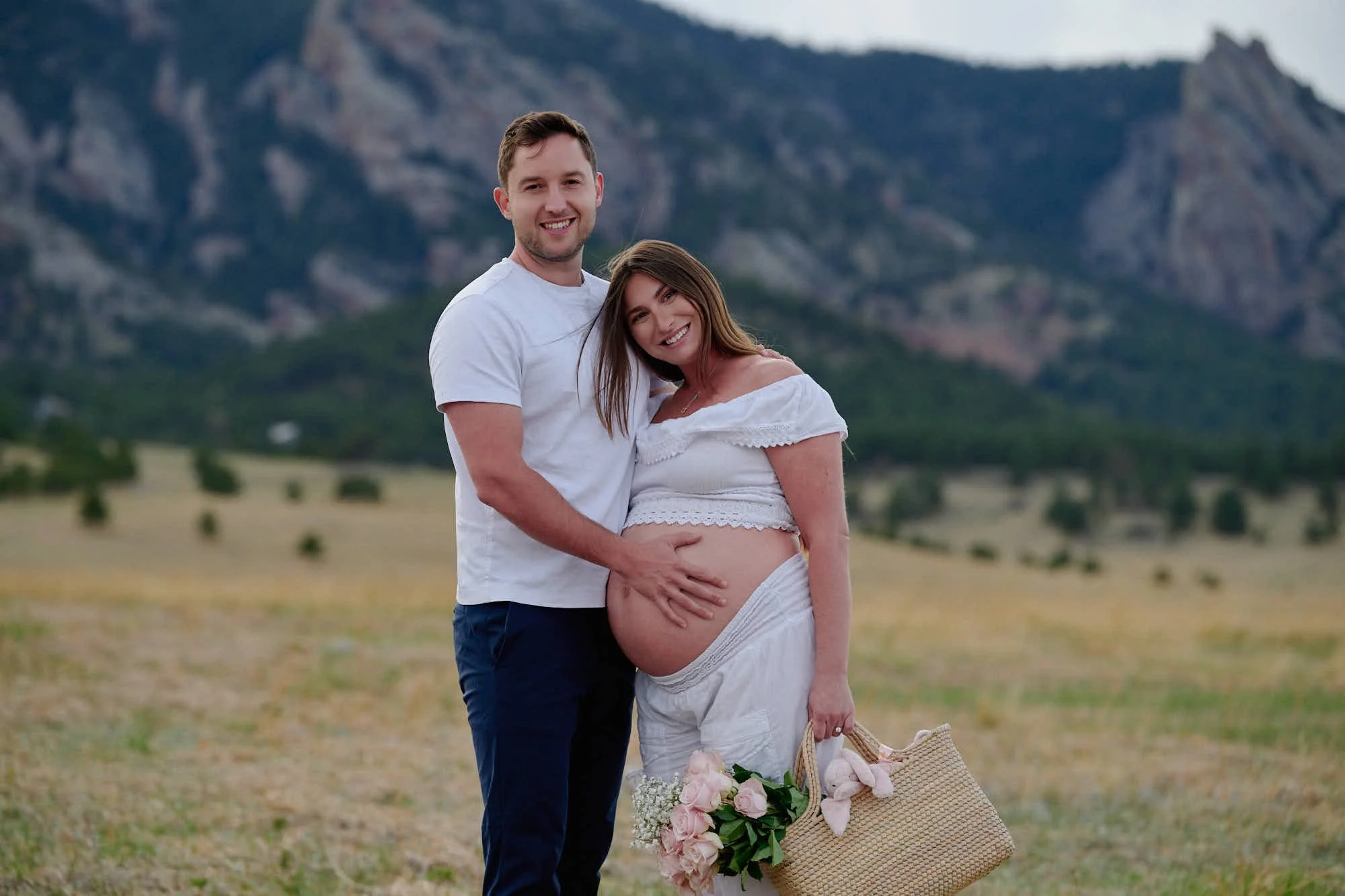 Expecting parents together in an open meadow at dusk, a woven basket of flowers at her feet.
