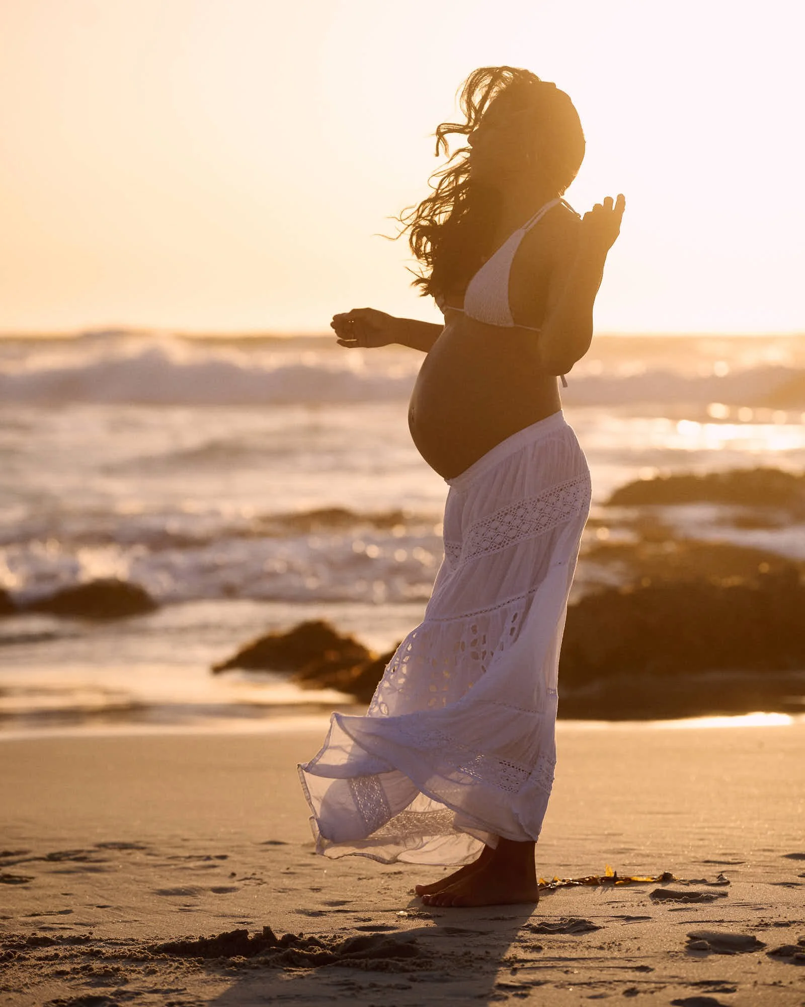 White lace skirt at golden hour, hands raised as the surf rolls up the Big Sur beach.
