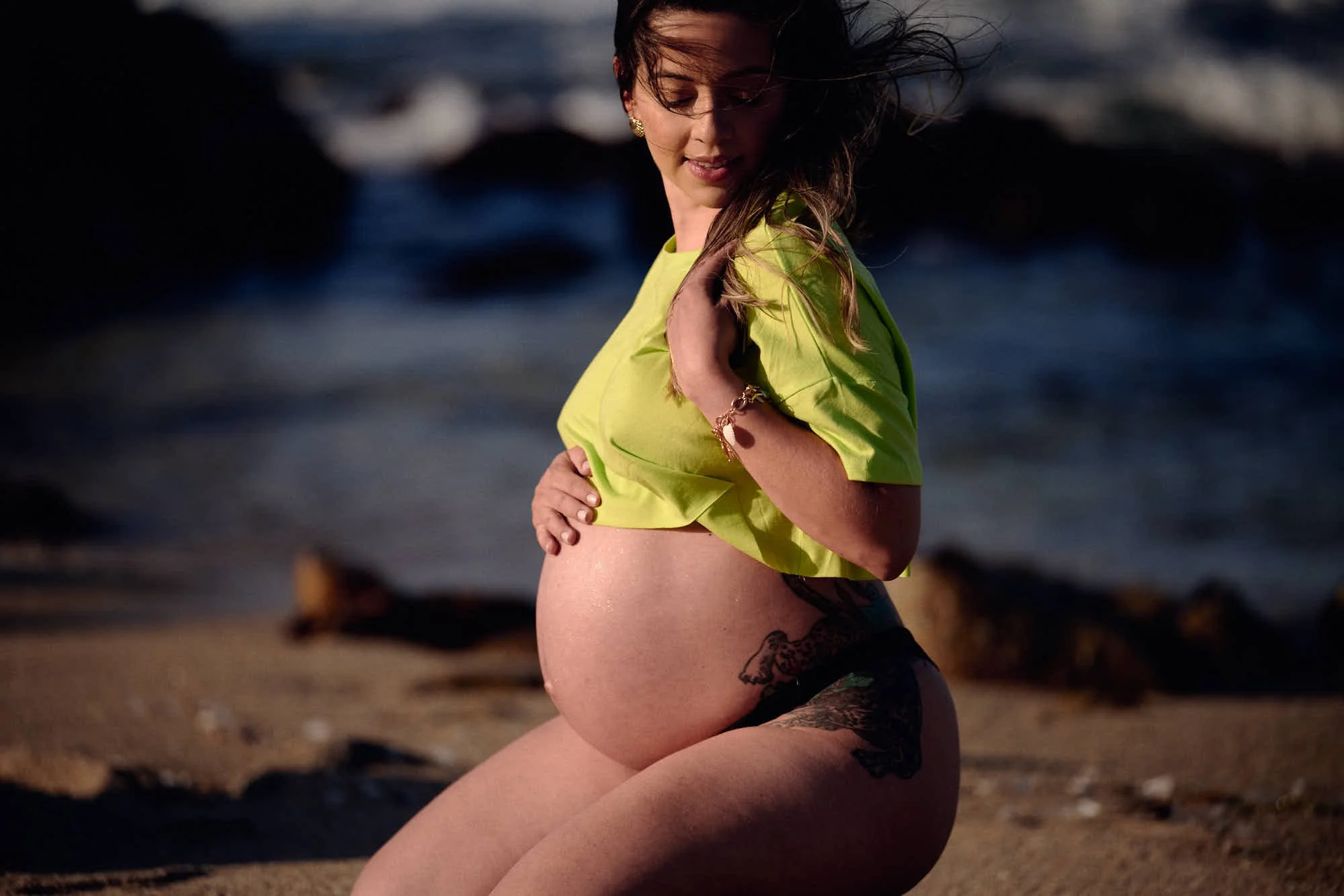 Lime green crop top and bare bump on a Big Sur beach, wind in her hair and dark rocks behind.