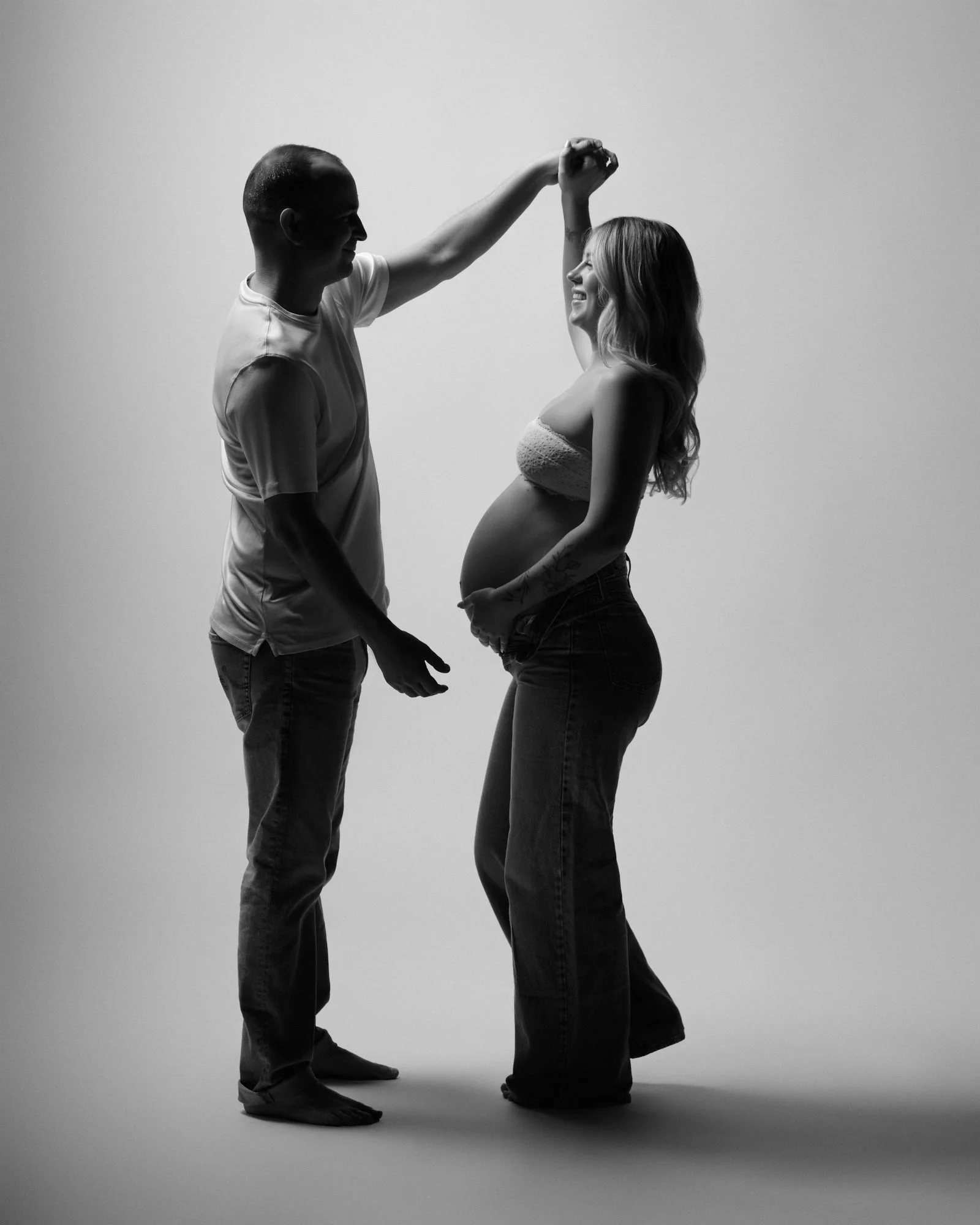 Mother-to-be twirls under her partner's arm mid-dance, laughing in moody light.