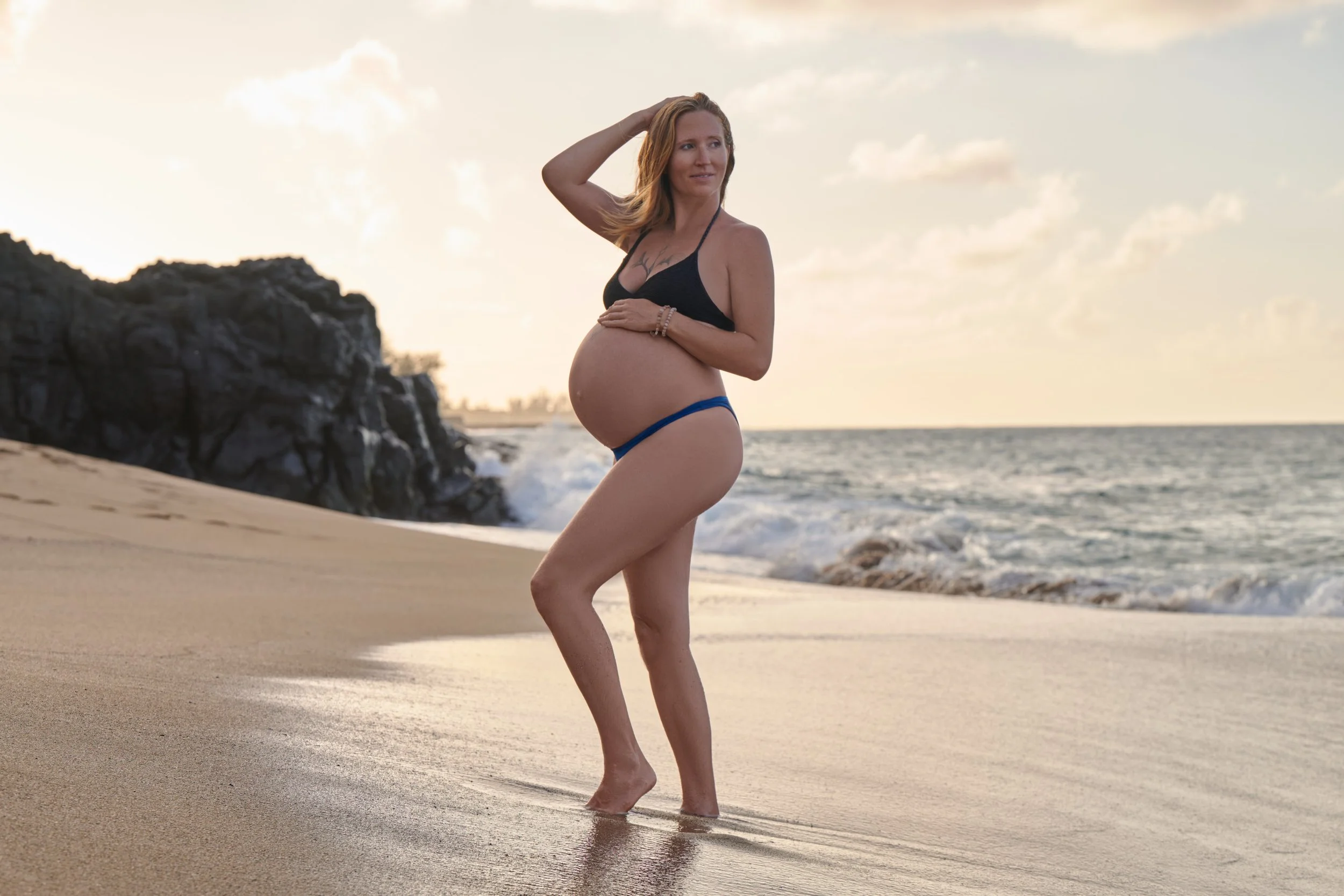 Black bikini top and blue bottoms on a Kauai beach, hand in her hair, lava rock and surf behind.