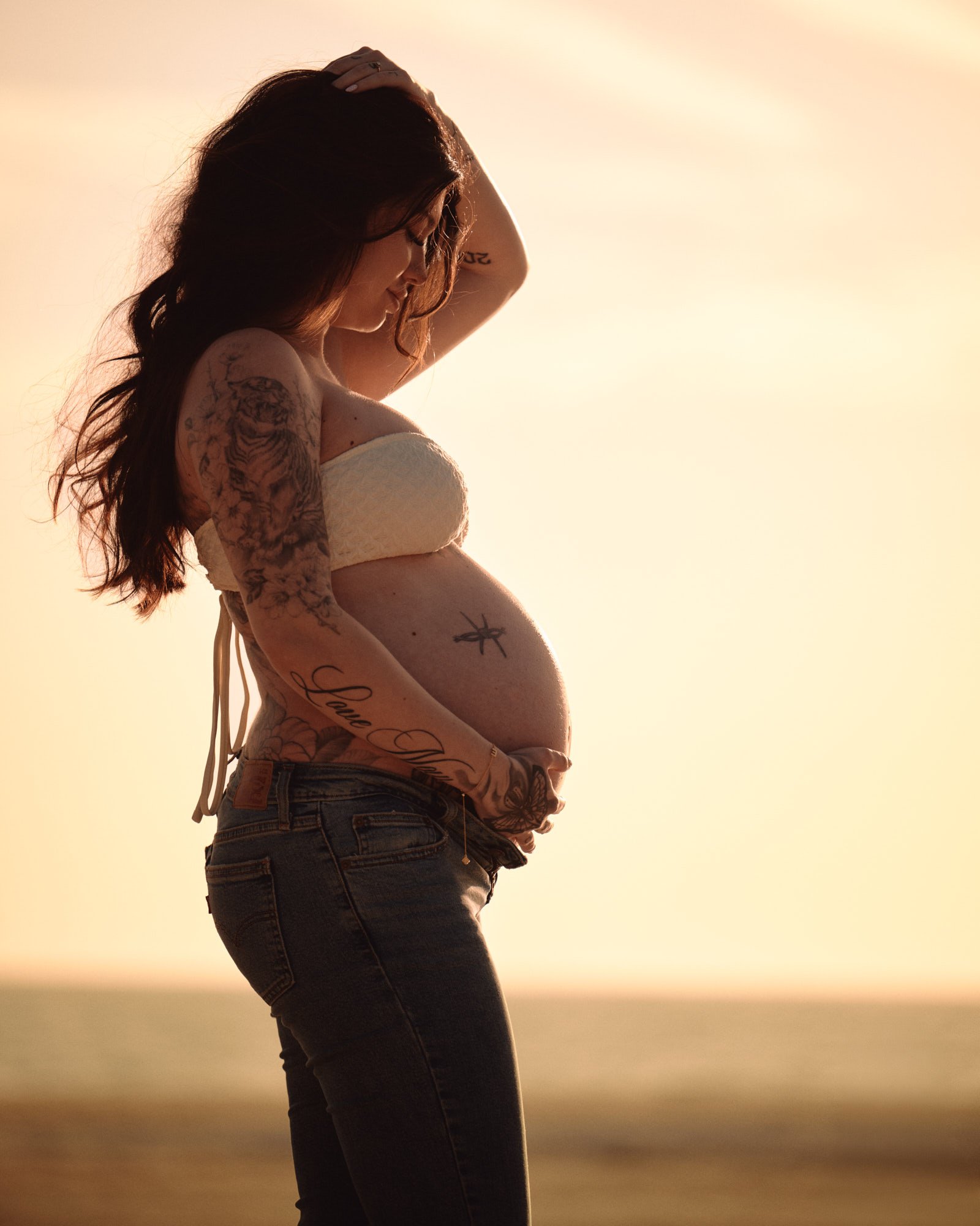 Hand shading her eyes at sunset, jeans and a white bandeau, warm light across the bump.