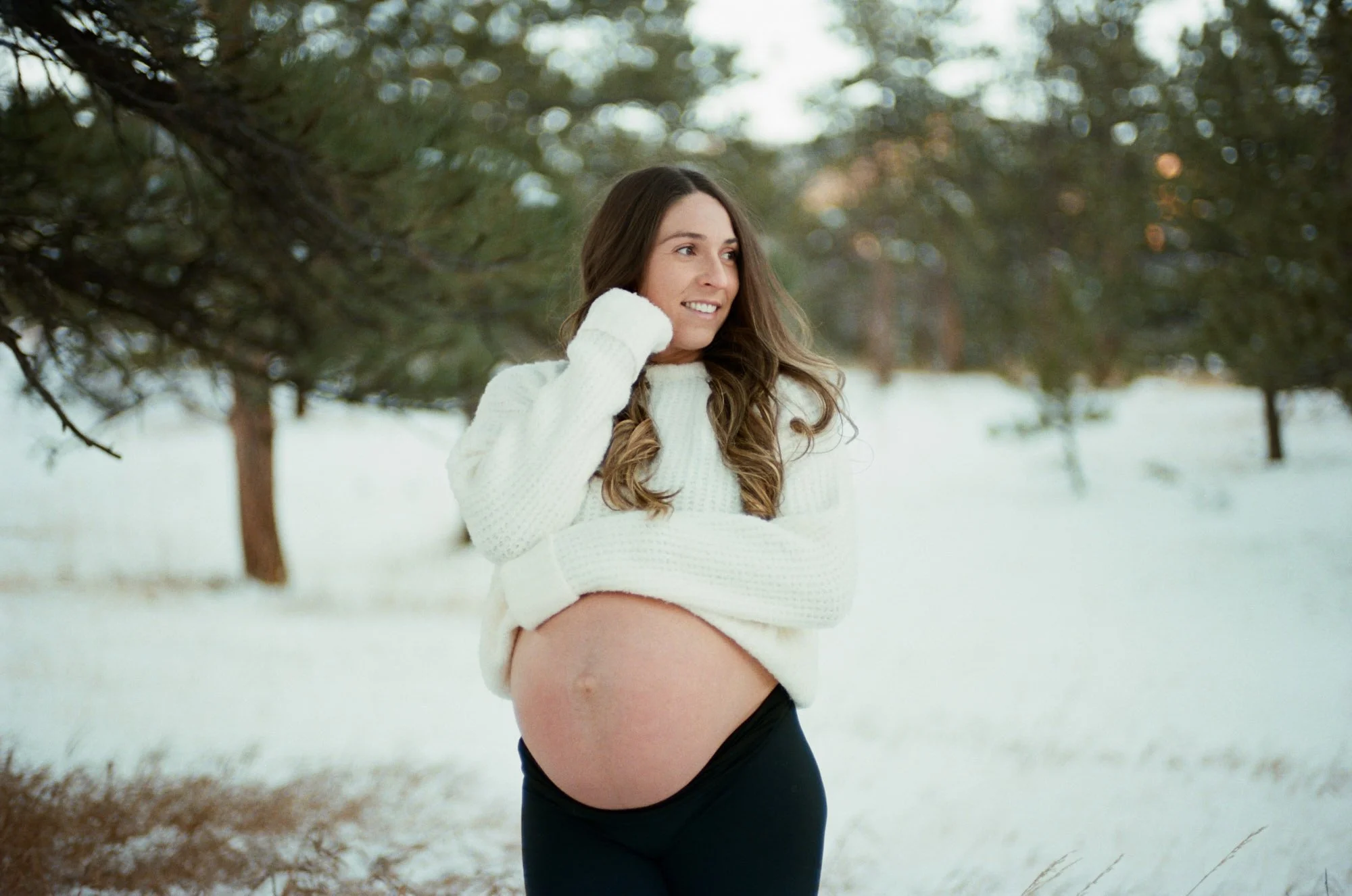 Cream sweater lifted over the bare bump in the snow, backlit among evergreens, shot on film.