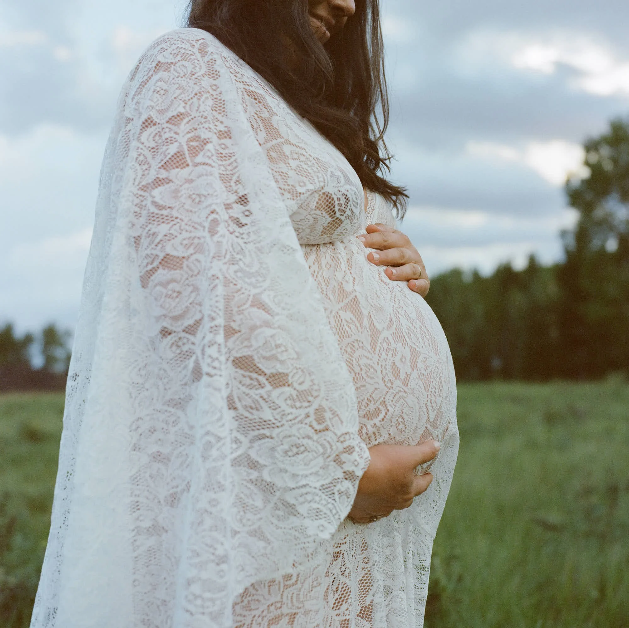 White lace sleeves and hands on the bump, meadow and pines soft behind on film.