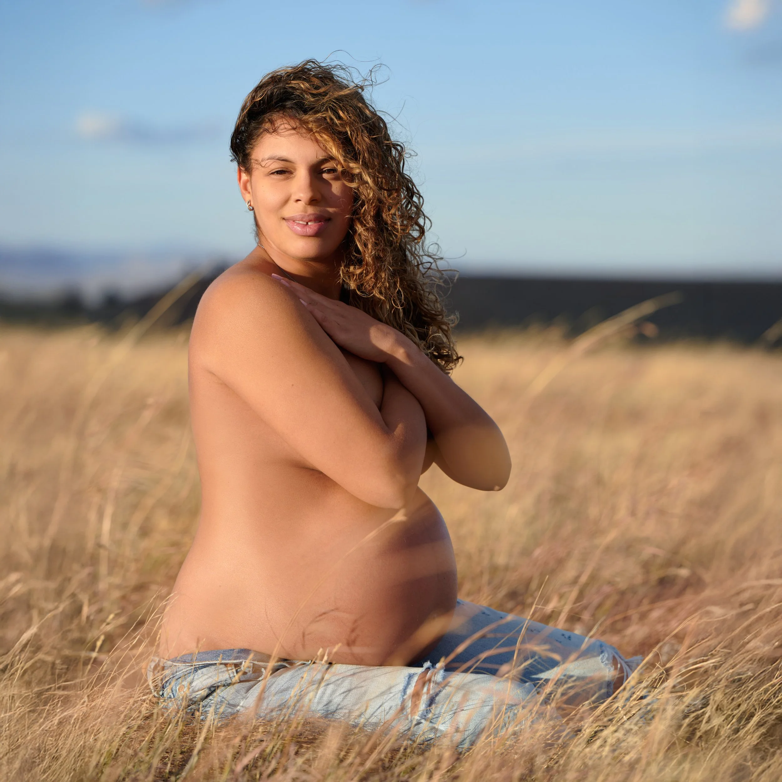 Seated in winter prairie grass, arms crossed softly, curls catching the cold sunlight.