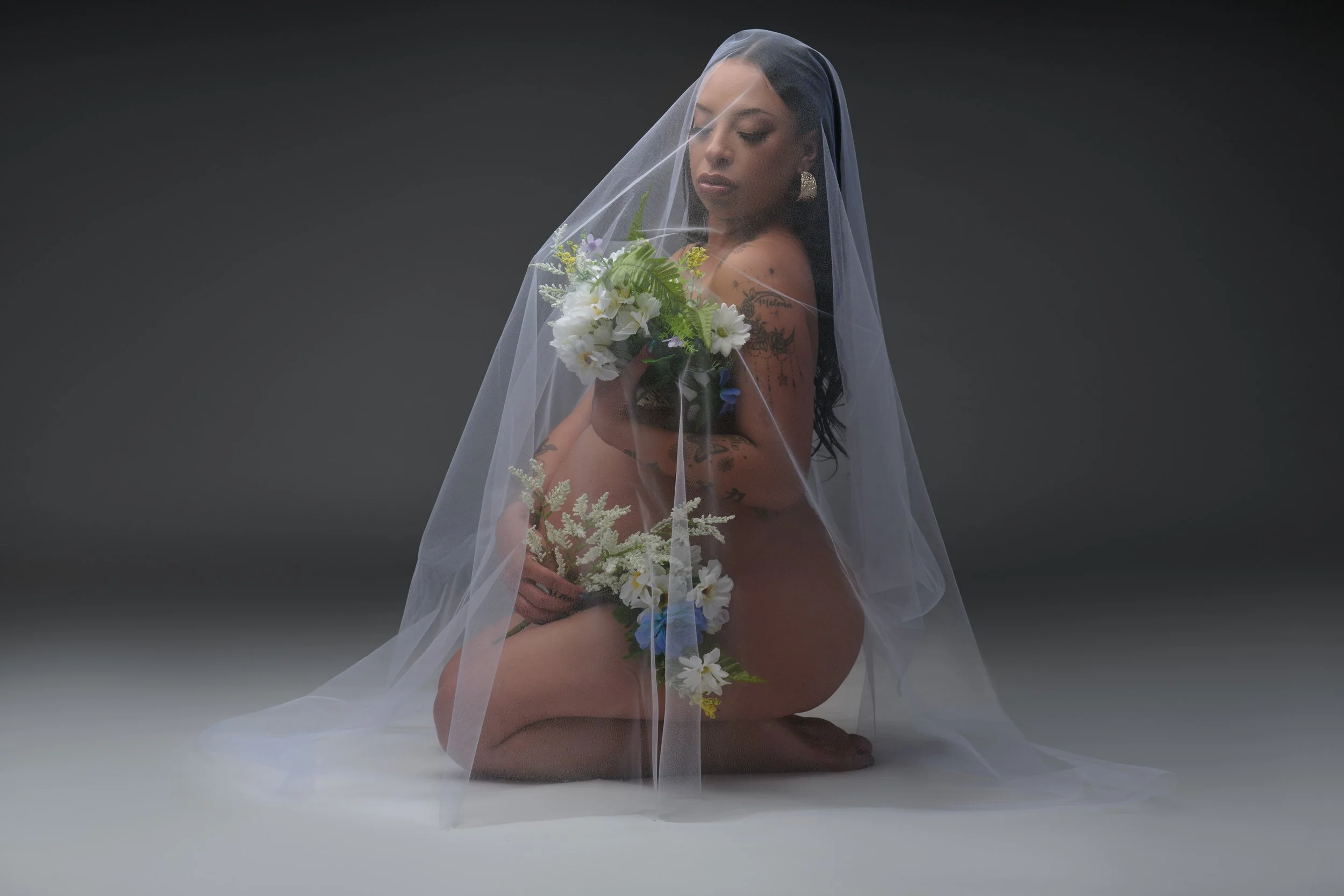 Kneeling under a long tulle veil with a daisy bouquet, lit soft at a Denver studio maternity session.