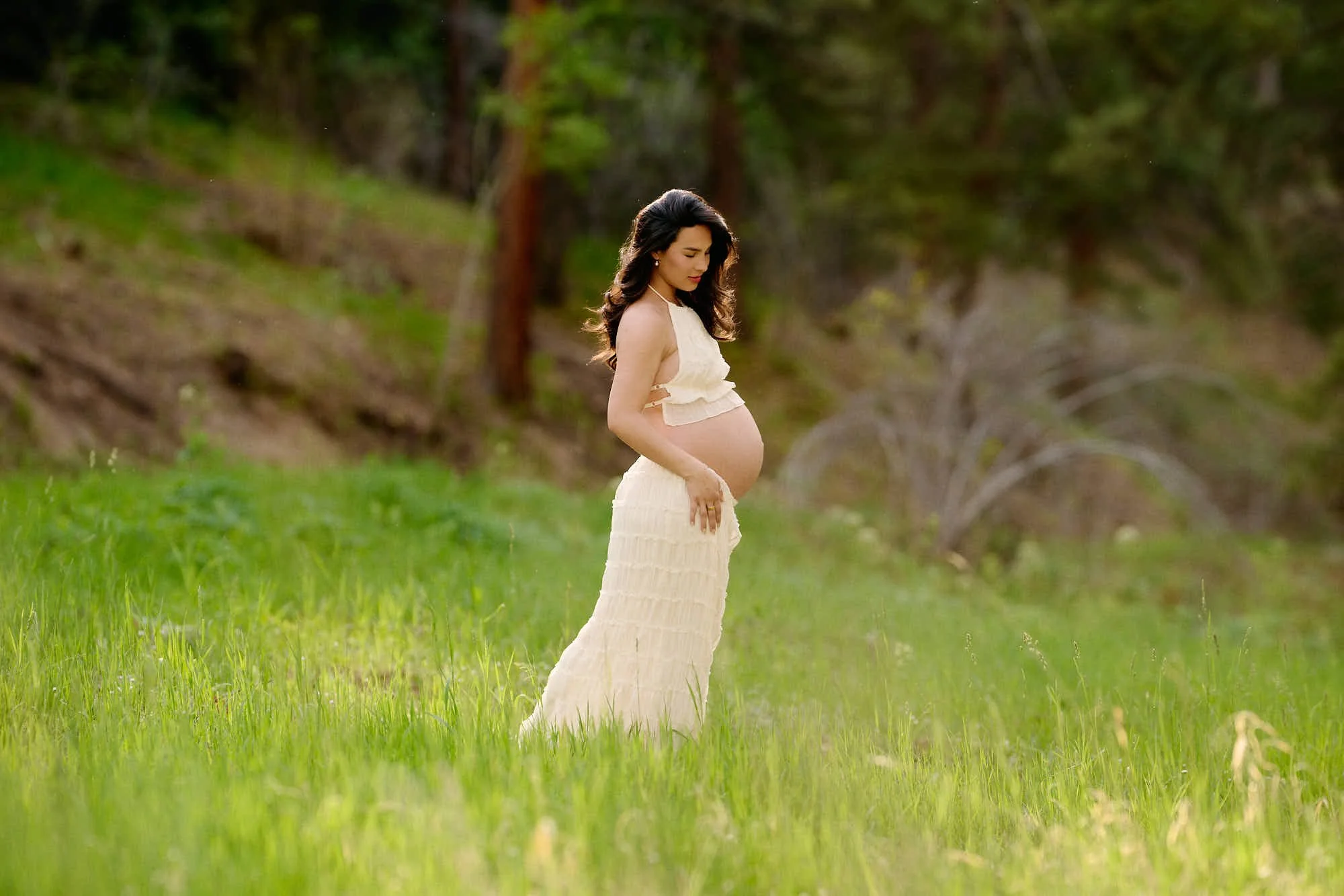 Outdoor maternity pose in a cream two-piece gown, standing in tall meadow grass below the pines.