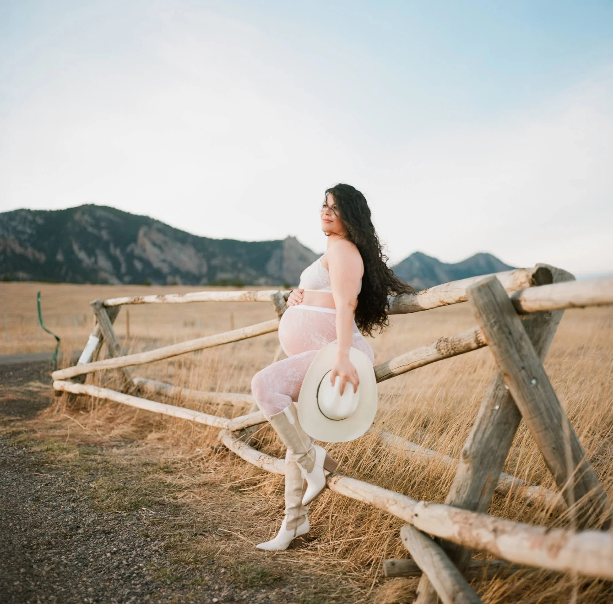 Pale slip dress and white boots against a weathered fence, straw hat held at her side.
