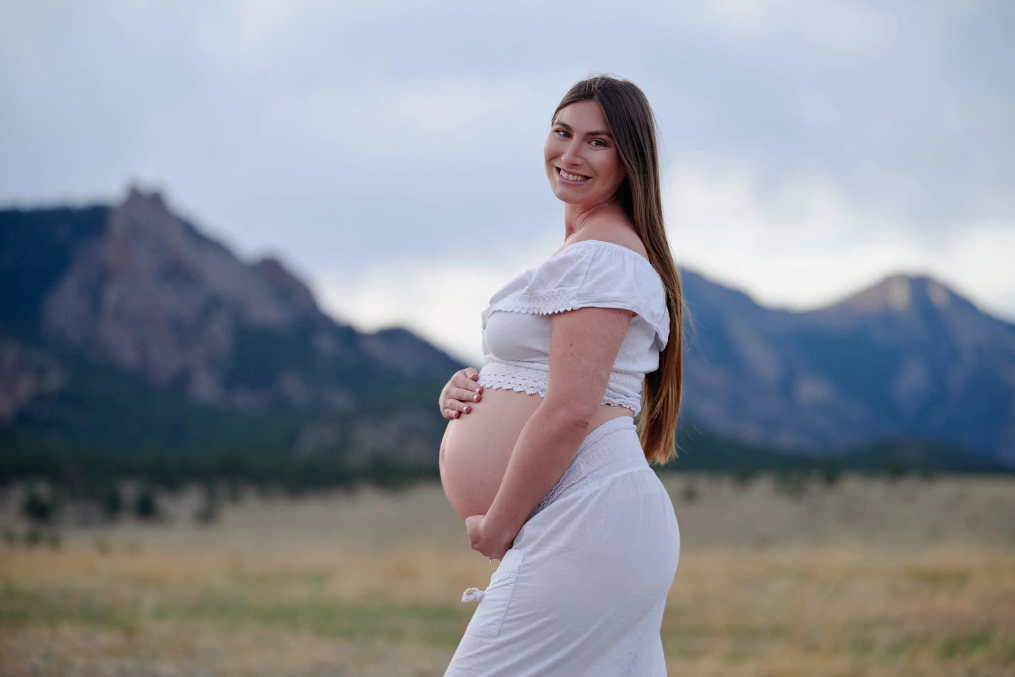 Mother to be in a white off-shoulder gown, hands under the bump, mountains soft behind her.