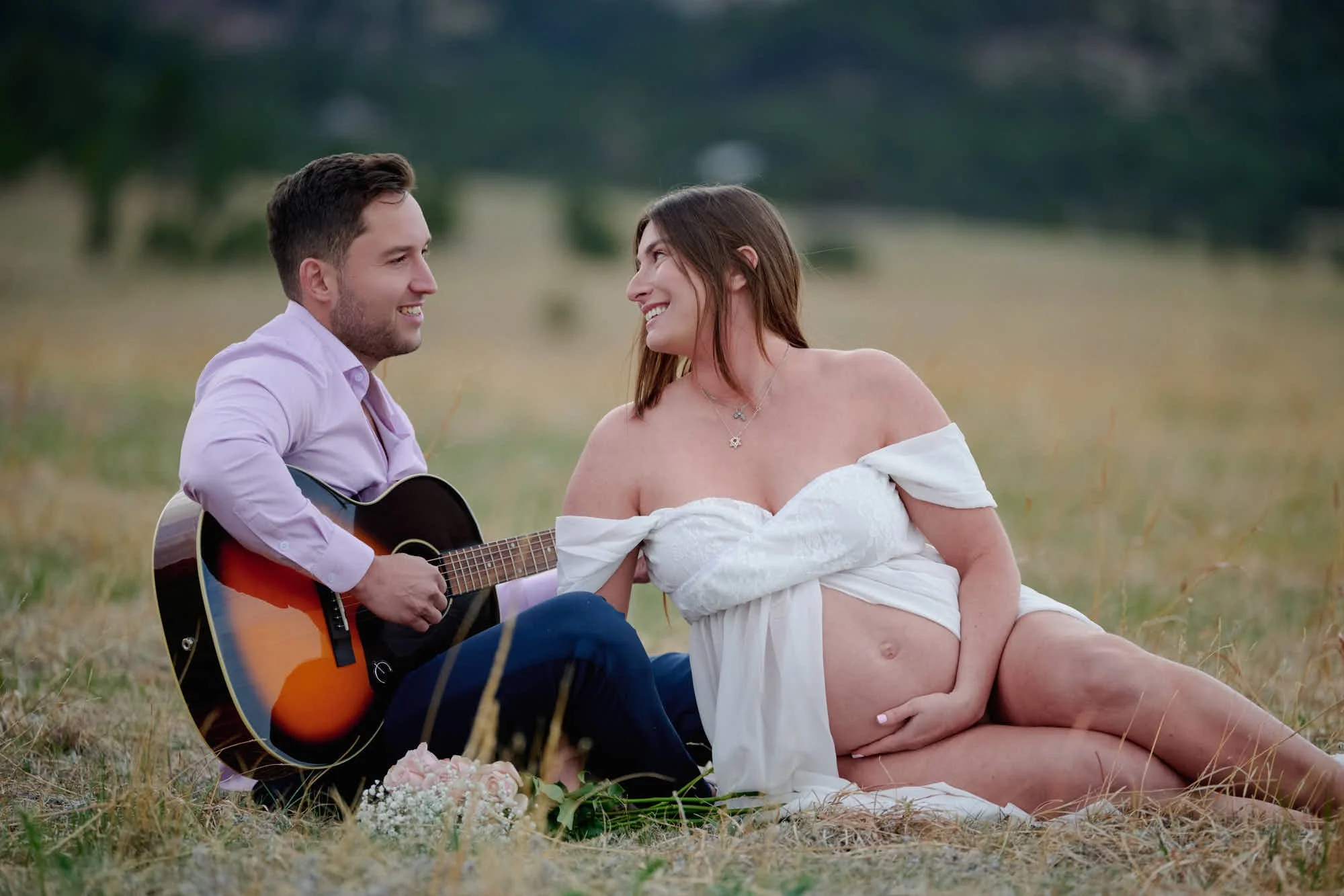 Partner plays acoustic guitar in the grass beside the mother-to-be during an early spring session.