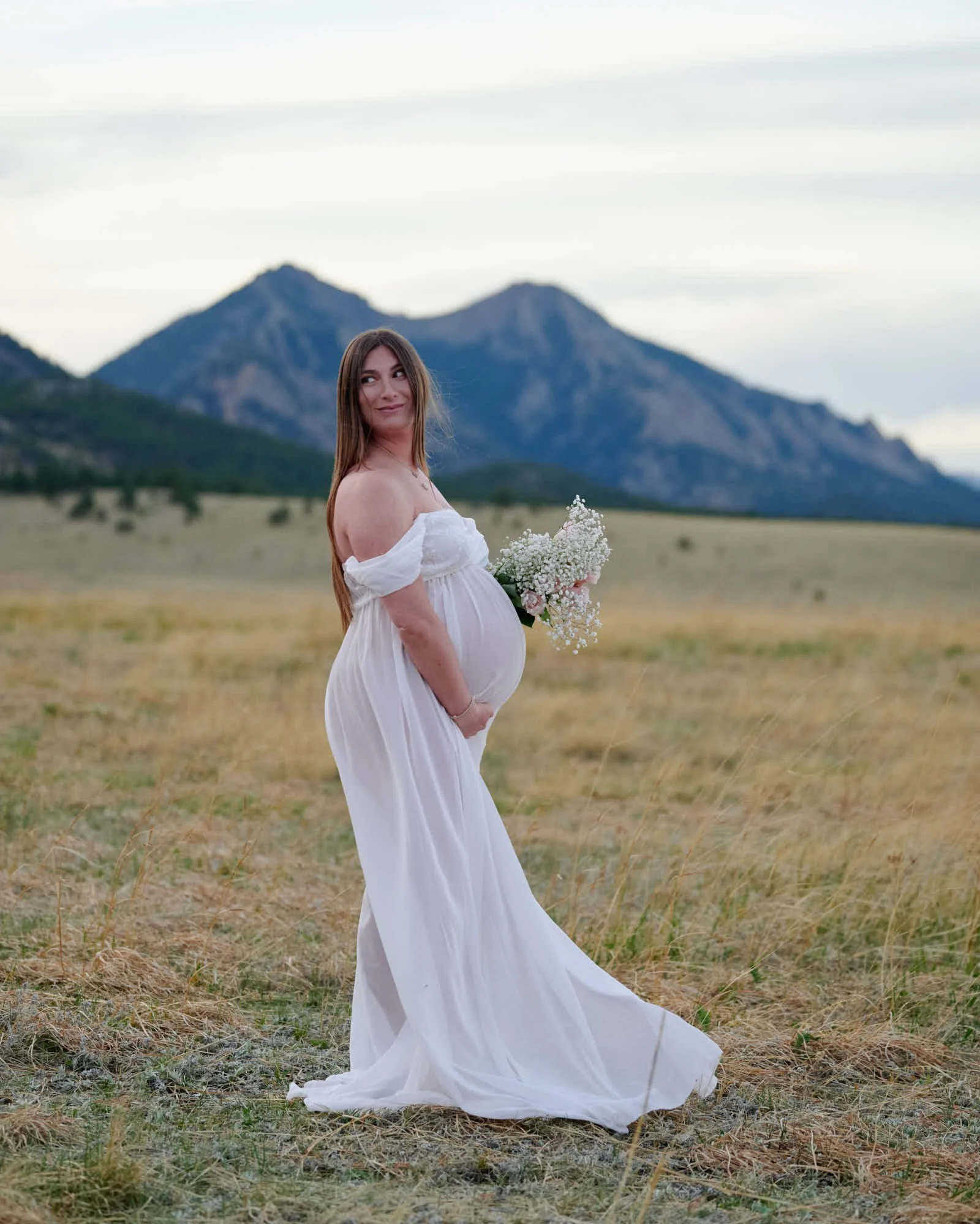 Full length spring maternity portrait, white flowing gown and a bouquet in open Colorado terrain.