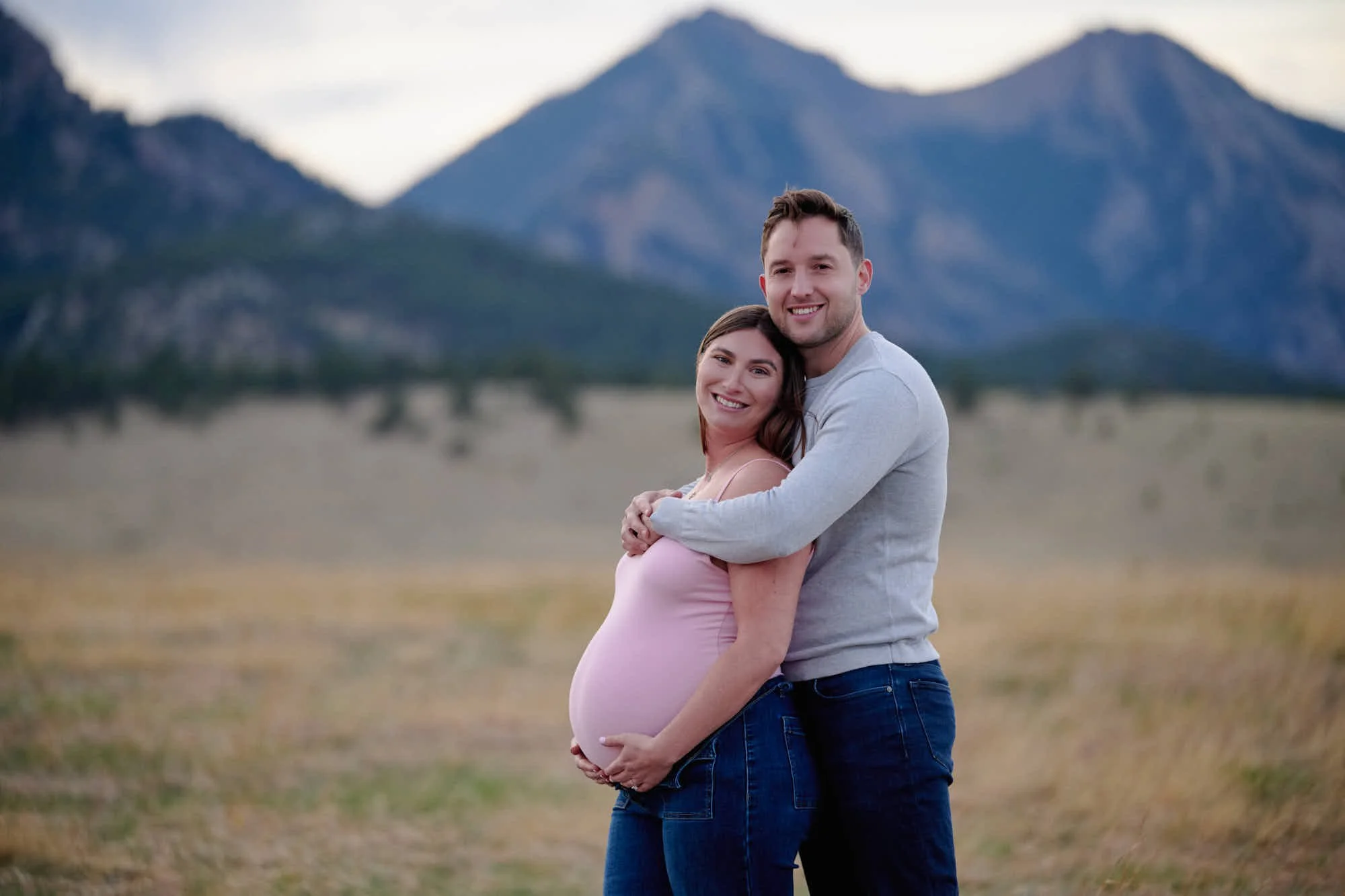 Partner wraps both arms around the bump in a pink dress, cool spring light across the meadow.