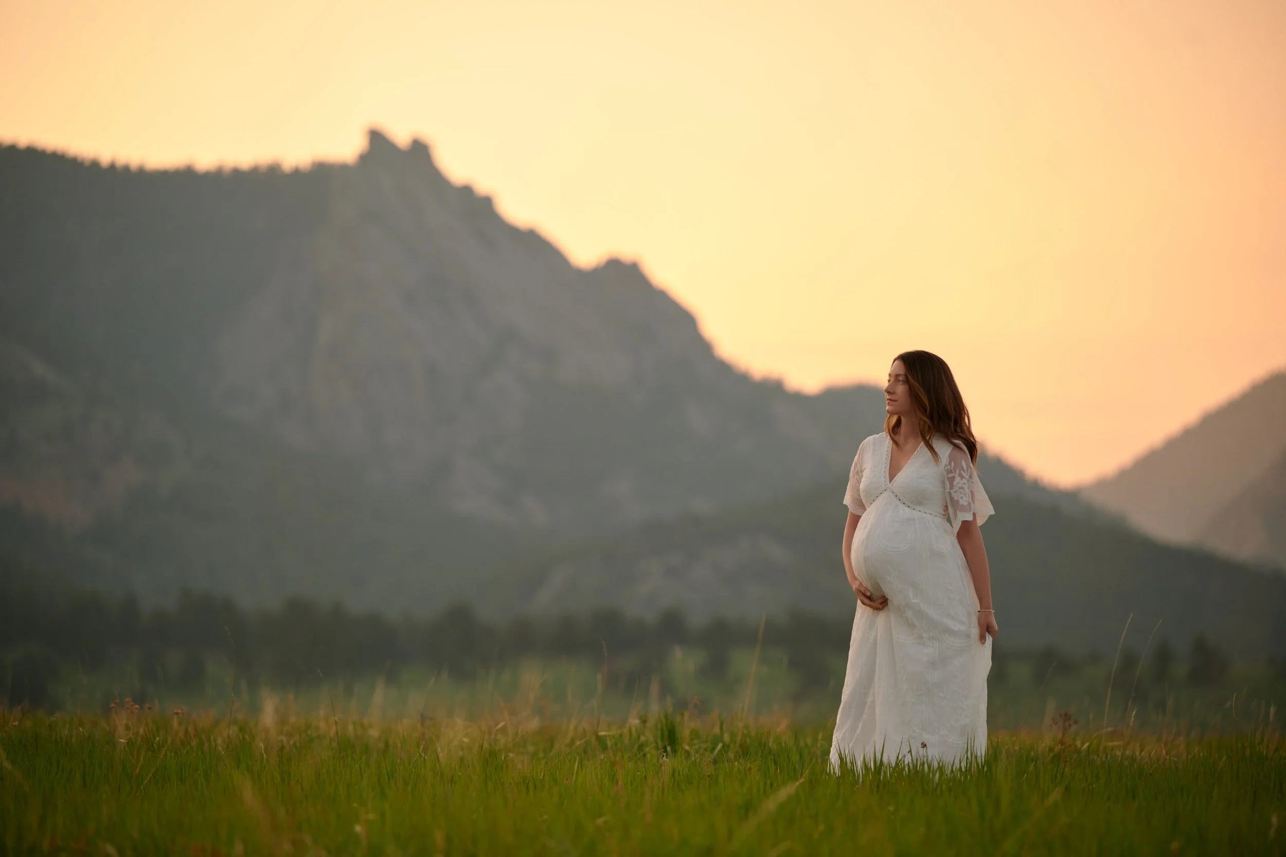 Alone in a wide golden field at sunset, mountain silhouettes stacked behind her white dress.