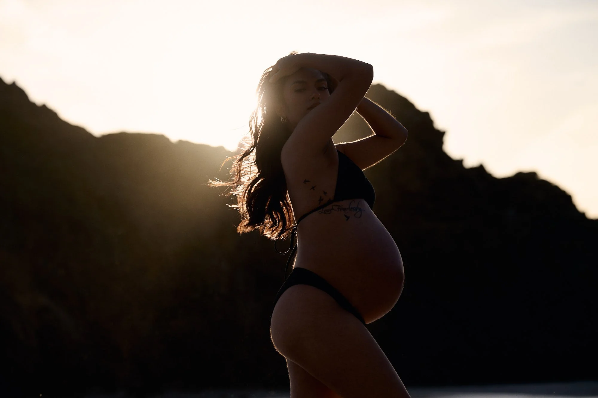 Sun-flared maternity portrait at sunset, hand raised to in hair in a black two-piece.