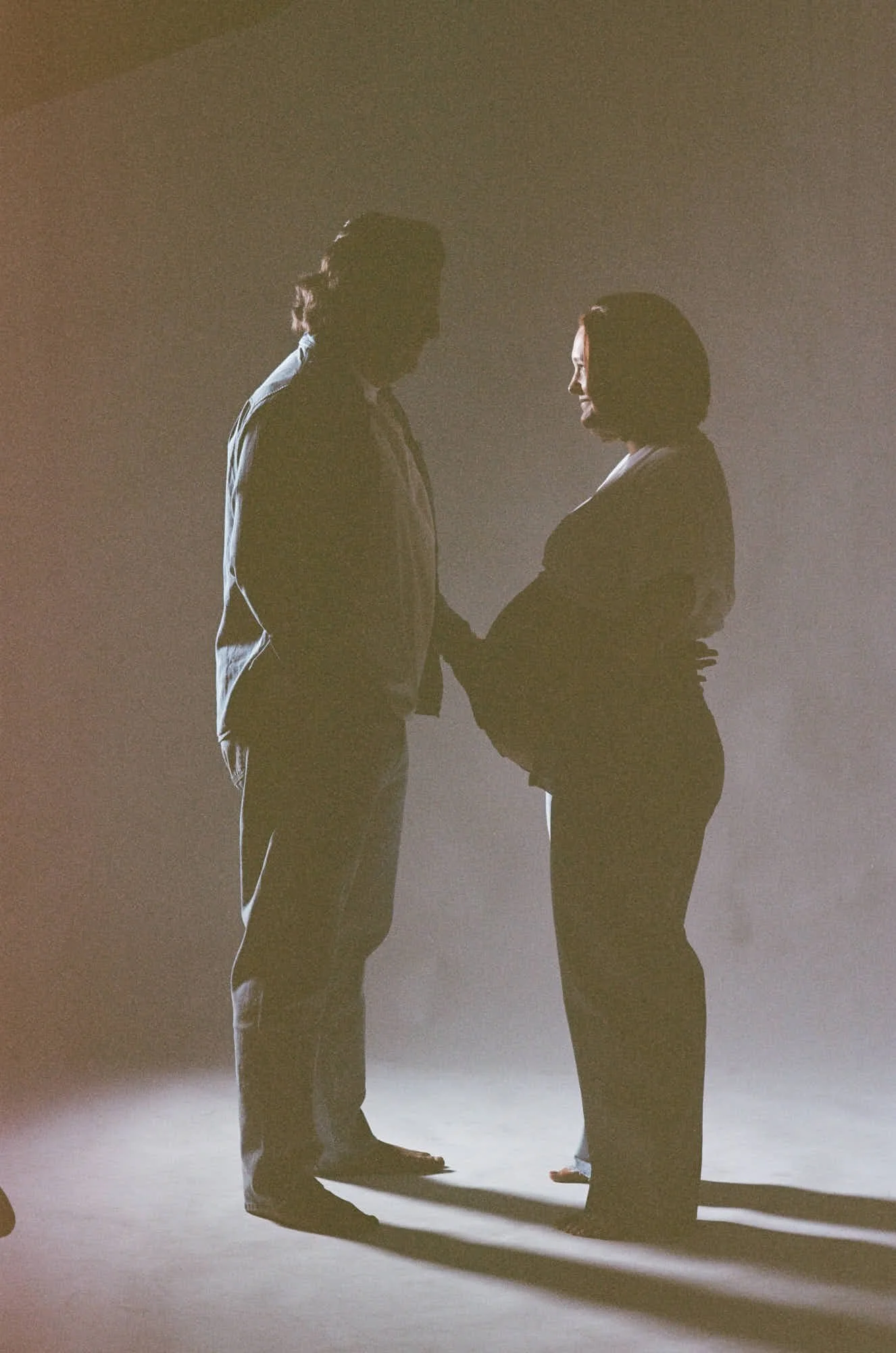 Two silhouettes facing each other in hard warm light, her bump between them and shadows on the wall.