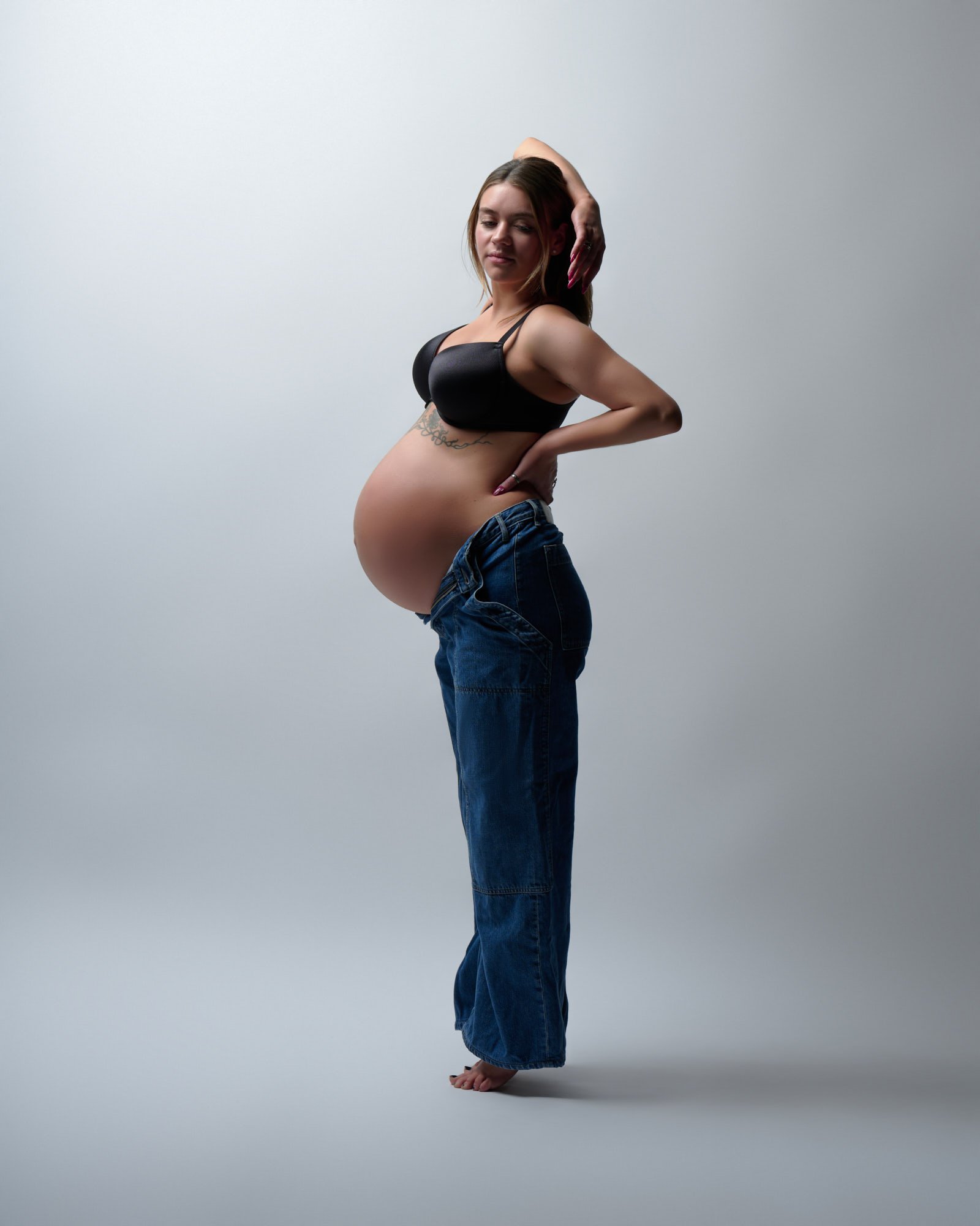 Arm arched overhead in unzipped jeans, bare bump in profile.