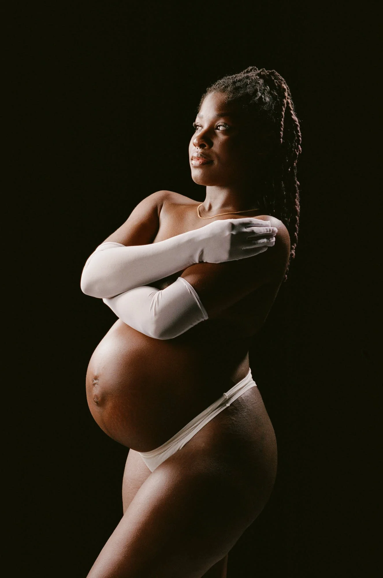 Long white gloves crossed over the chest, braids and bare shoulders lit against a dark room.