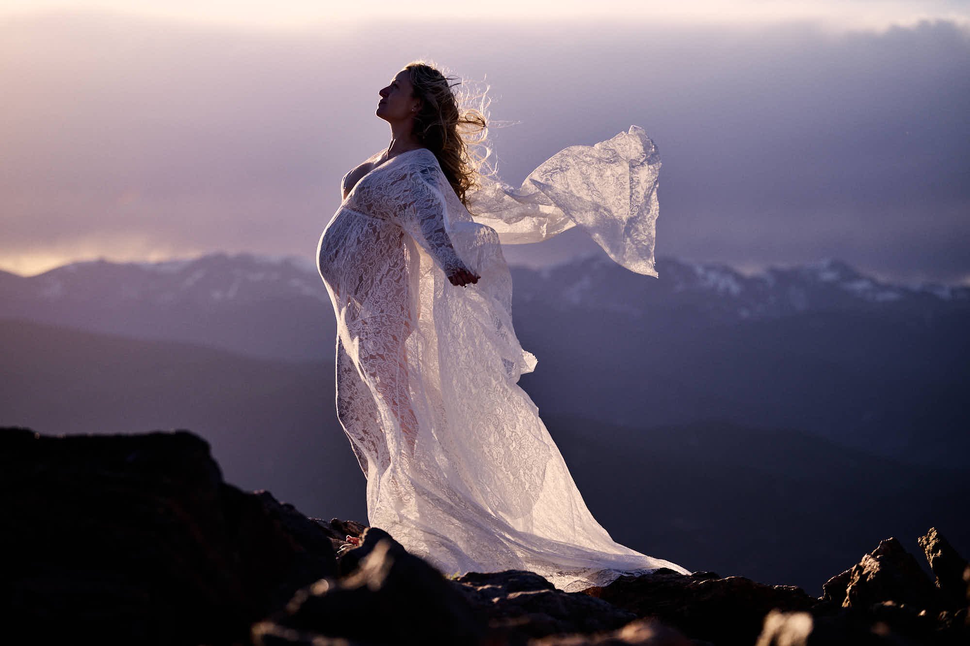 Adventure maternity at altitude: sheer lace gown swept by wind on a rocky summit at sunset, by Ethan Herrold.