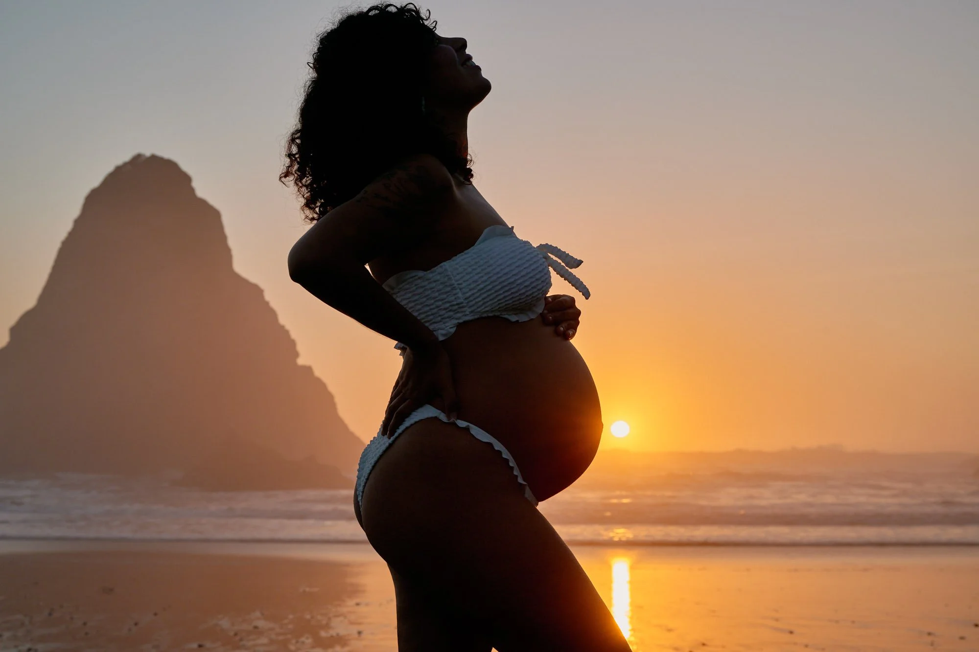 Silhouetted against the setting sun beside a sea stack, hands cradling the bump on the Oregon coast.