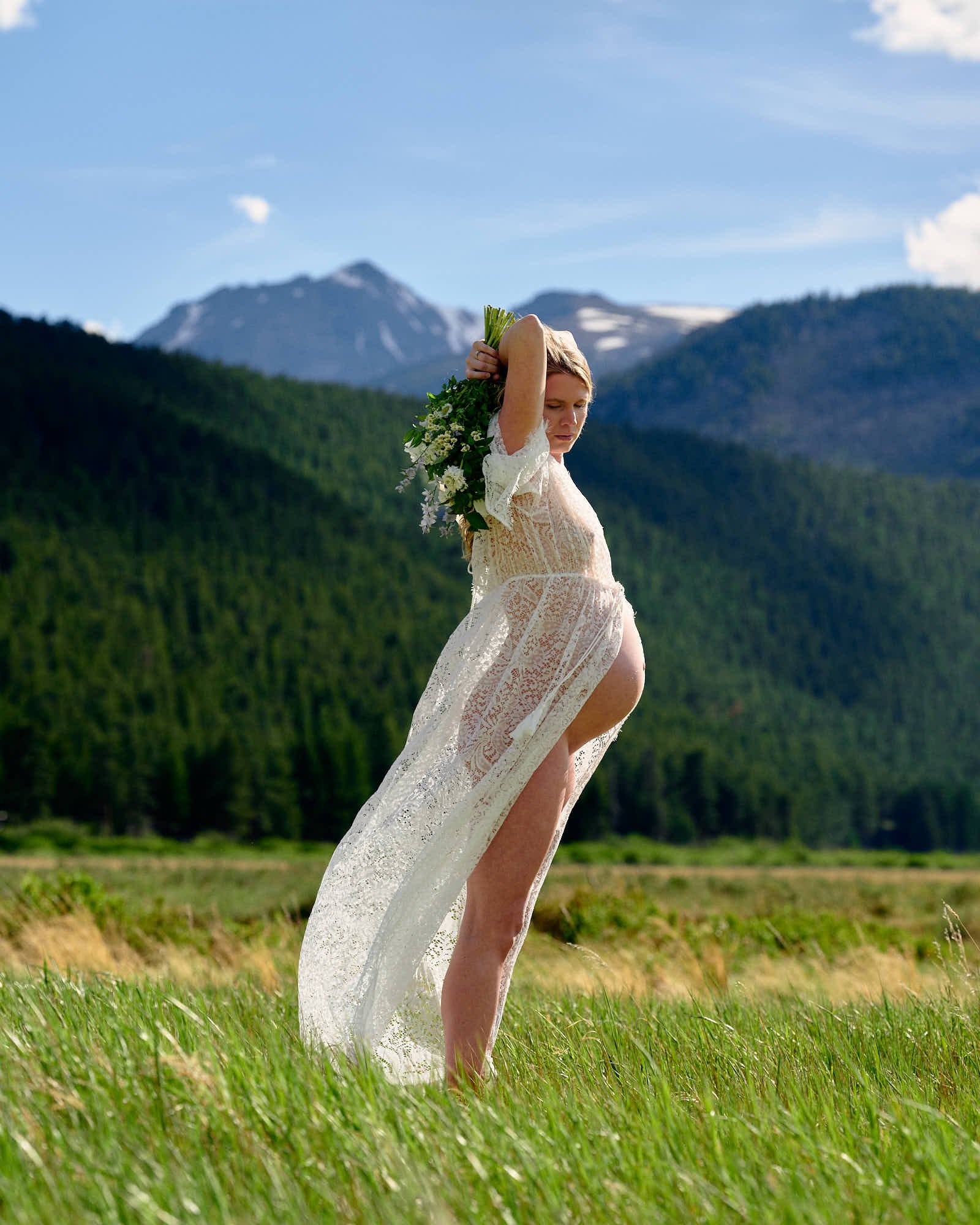 White lace gown and a raised bouquet in a mountain meadow below snow-streaked peaks.