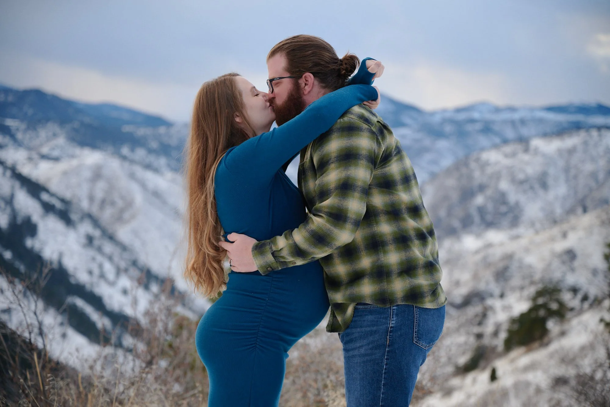 Couple kissing on a snowy slope, her in a long teal gown and him in a green flannel.