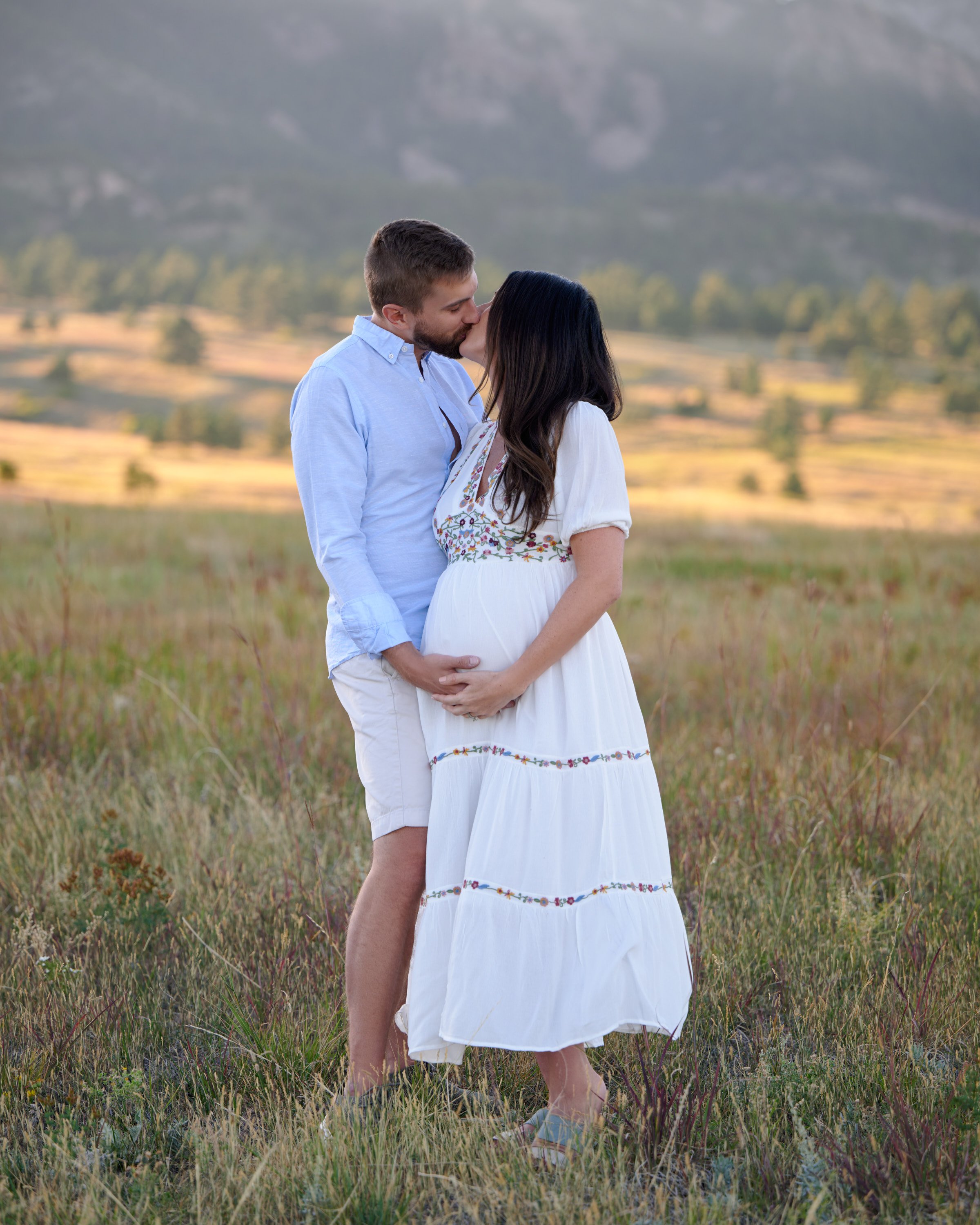 A kiss on the forehead in a golden valley meadow, evening alpenglow on the ridgeline.