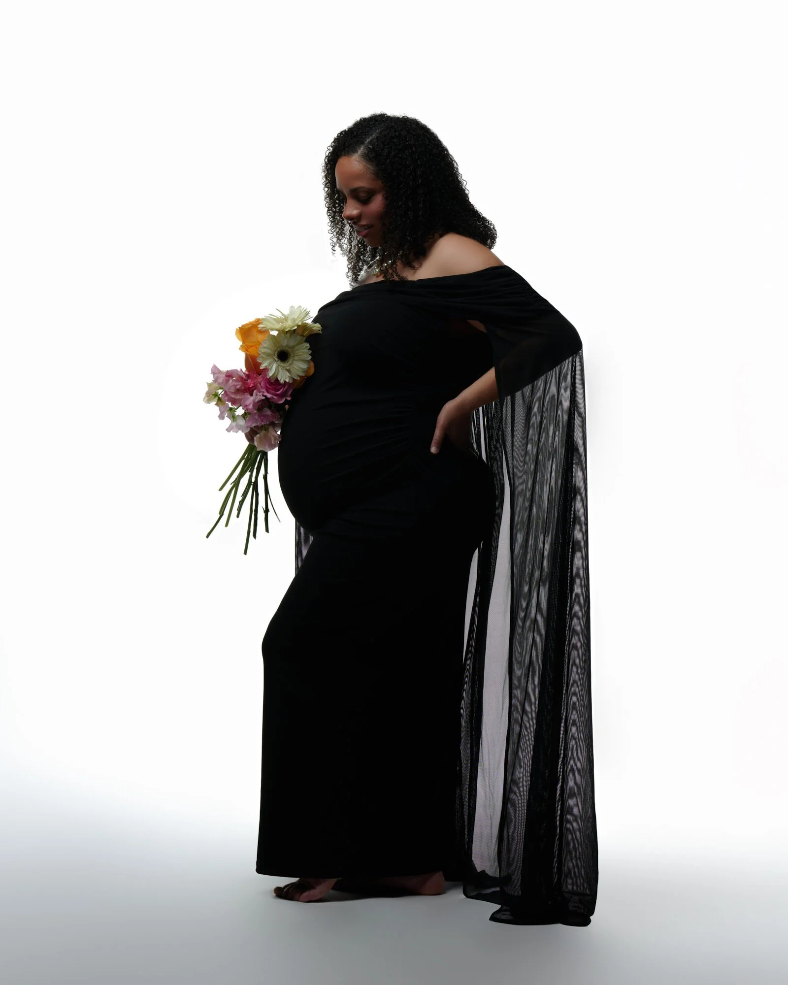 Black gown with sheer cape sleeves and a bouquet, glowing backlight behind.