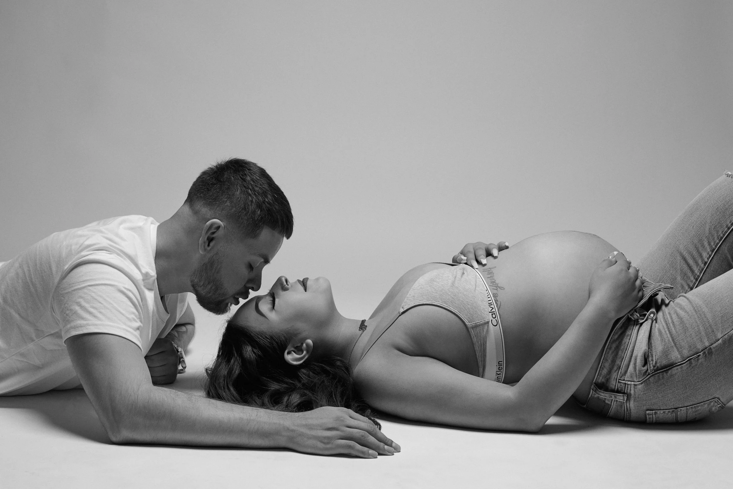 A couple lying on the floor facing each other, with their foreheads and noses touching. The woman is pregnant and wearing a tank top and jeans, while the man is wearing a t-shirt. The scene is captured in black and white.