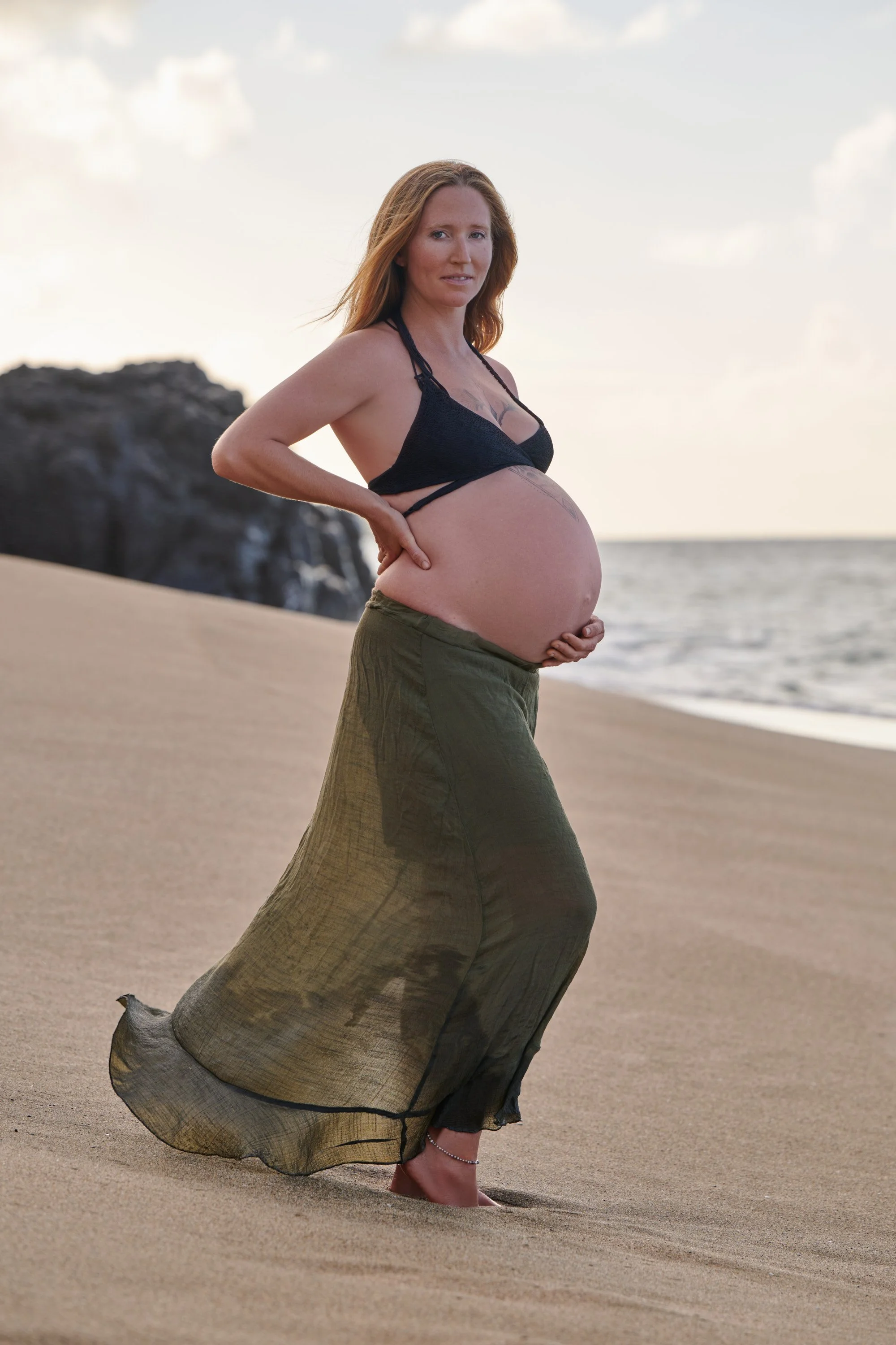 Olive skirt and black top on a Kauai beach, lava rock and surf behind her.