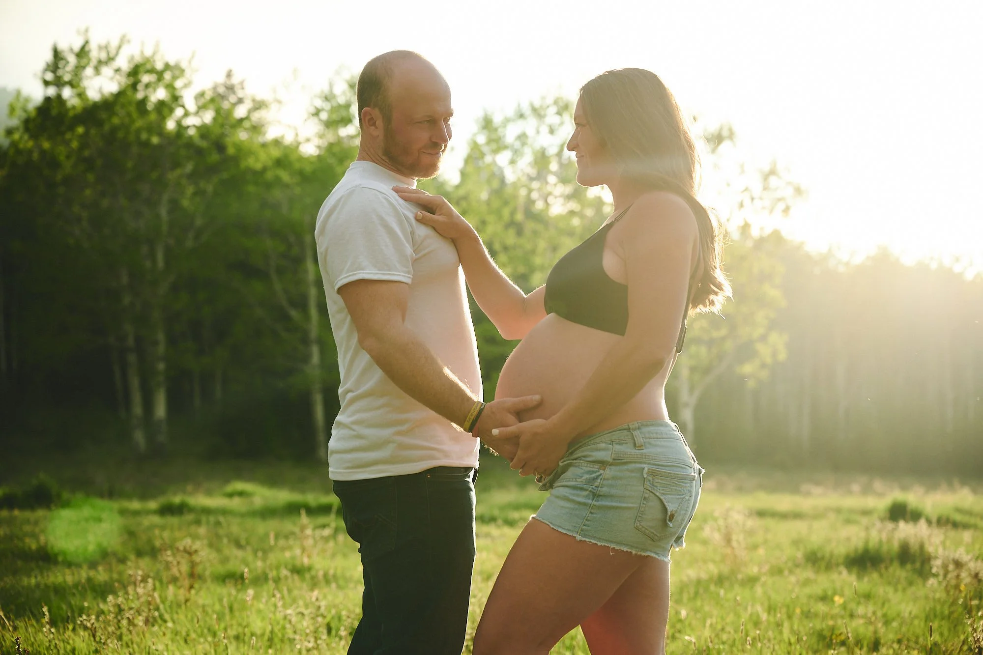 A pregnant woman and her partner stand in a grassy field with trees in the background, holding her belly and smiling at each other as the sun sets behind them.