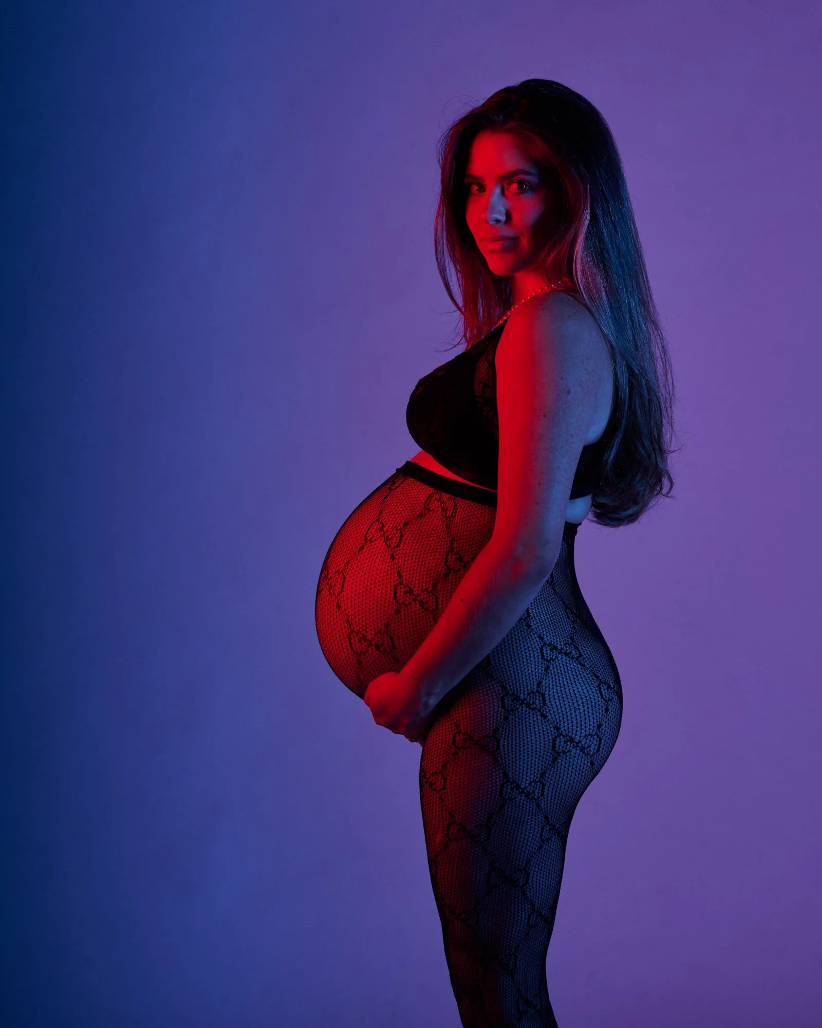 A woman with long hair in black lingerie and patterned fishnet tights stands against a purple and red gradient background, looking at the camera with a confident expression.