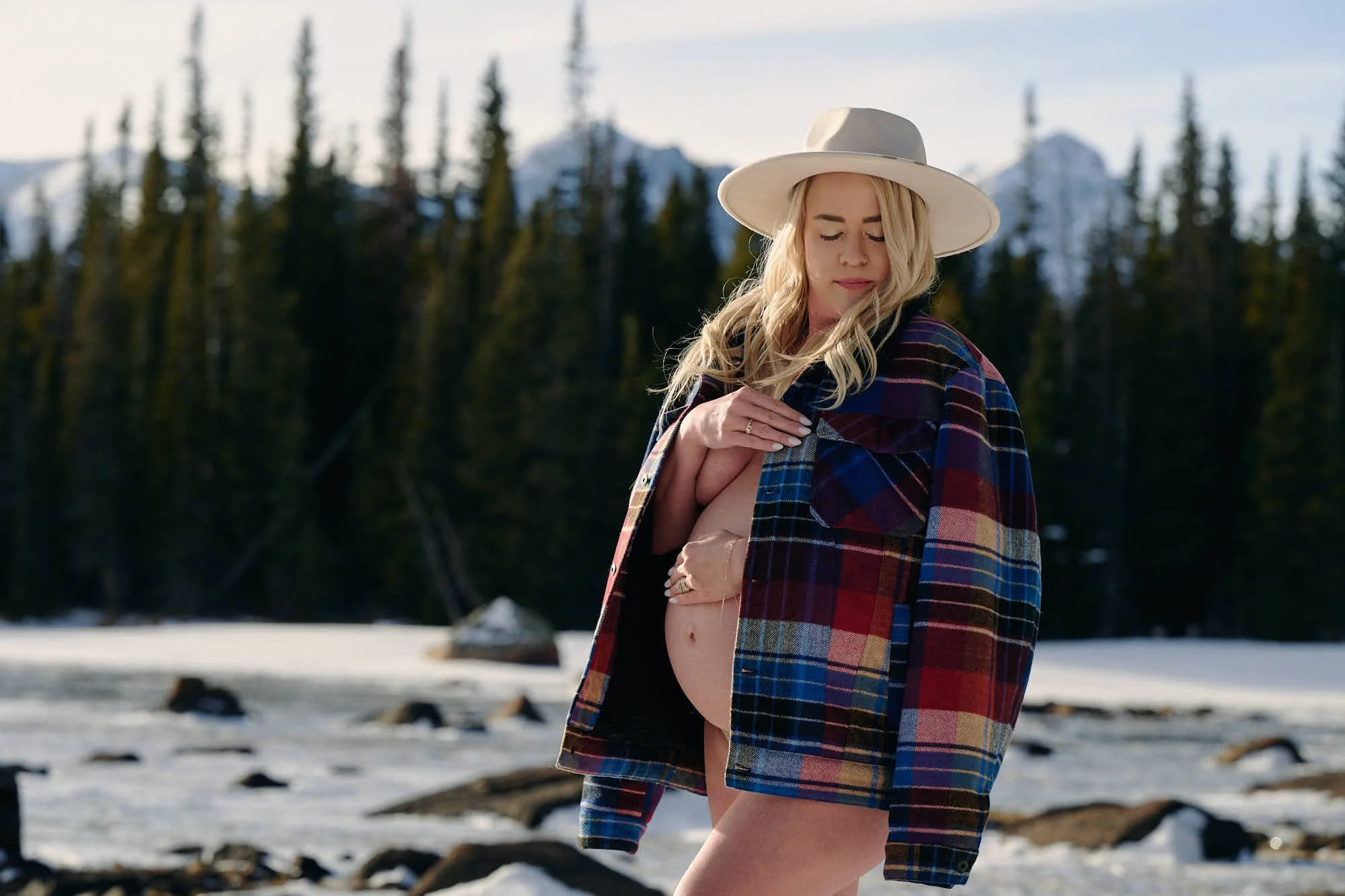 Wide-brim hat and an open plaid shacket over a bare bump on a frozen lakebed among spruce.