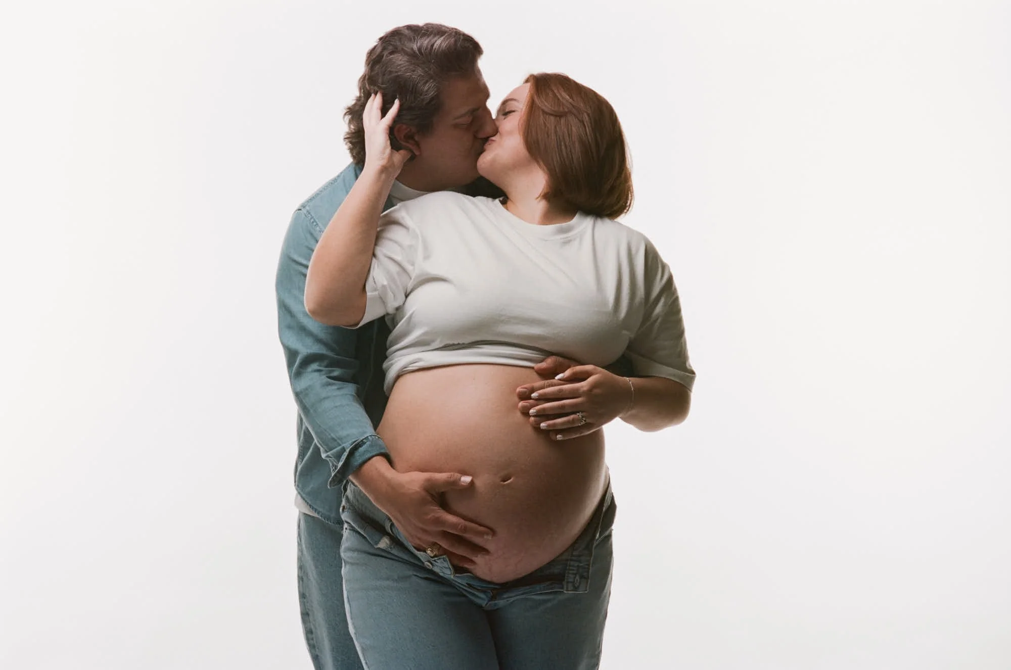 Expecting couple in matching gray tees kissing on a white studio set, shot on medium format film.