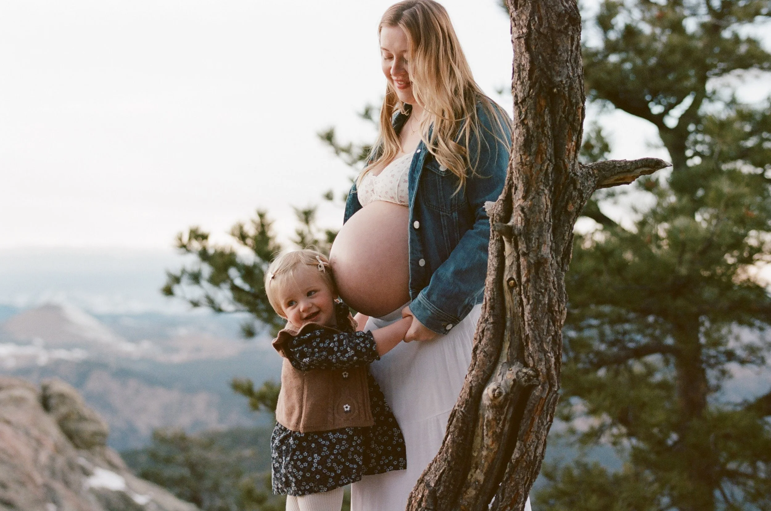 Mother and toddler beneath a wind-bent pine, canyon light open behind them on color film.