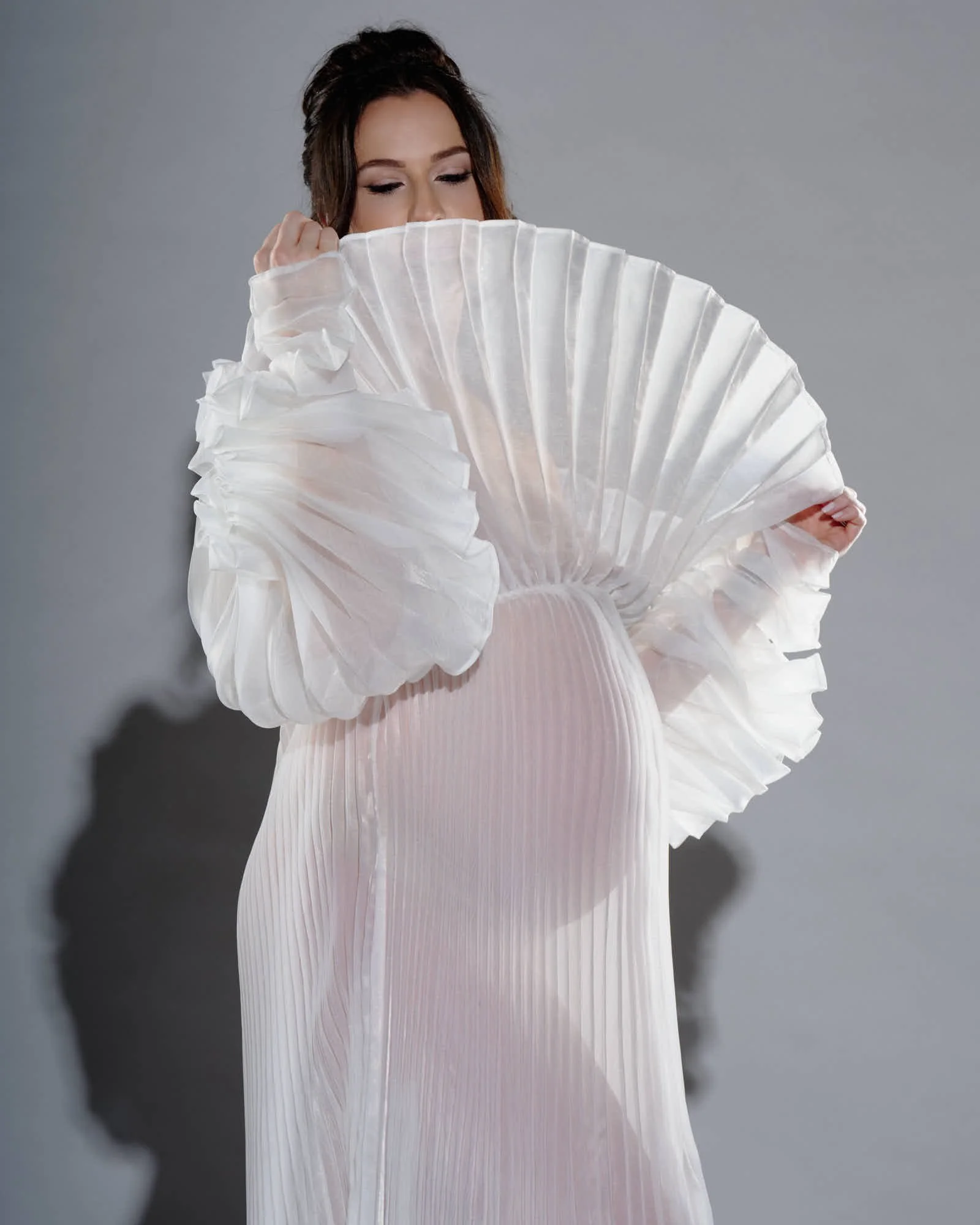 Pregnant woman behind a giant pleated fan collar, her bump visible through the sheer gown.