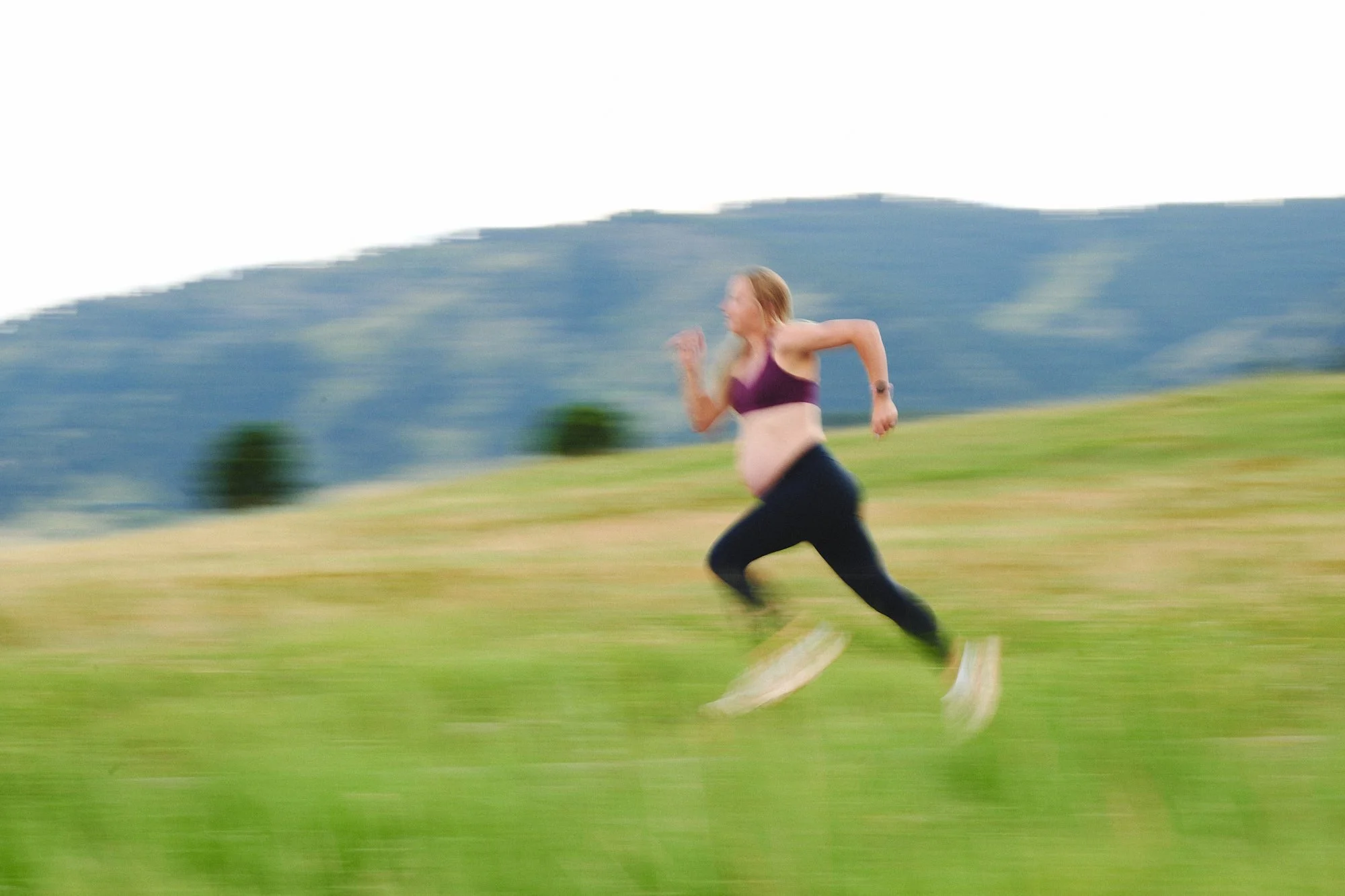 Pregnant runner blurring past a green Colorado hillside, stride long and unhurried.