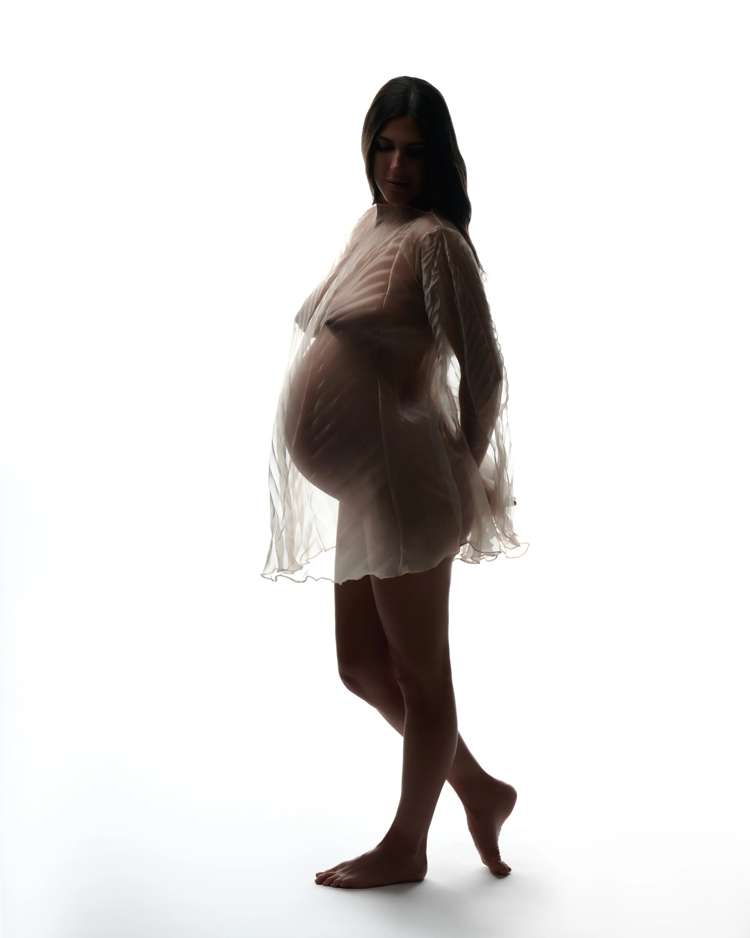Silhouette of a pregnant woman in a sheer dress standing barefoot against a white background.