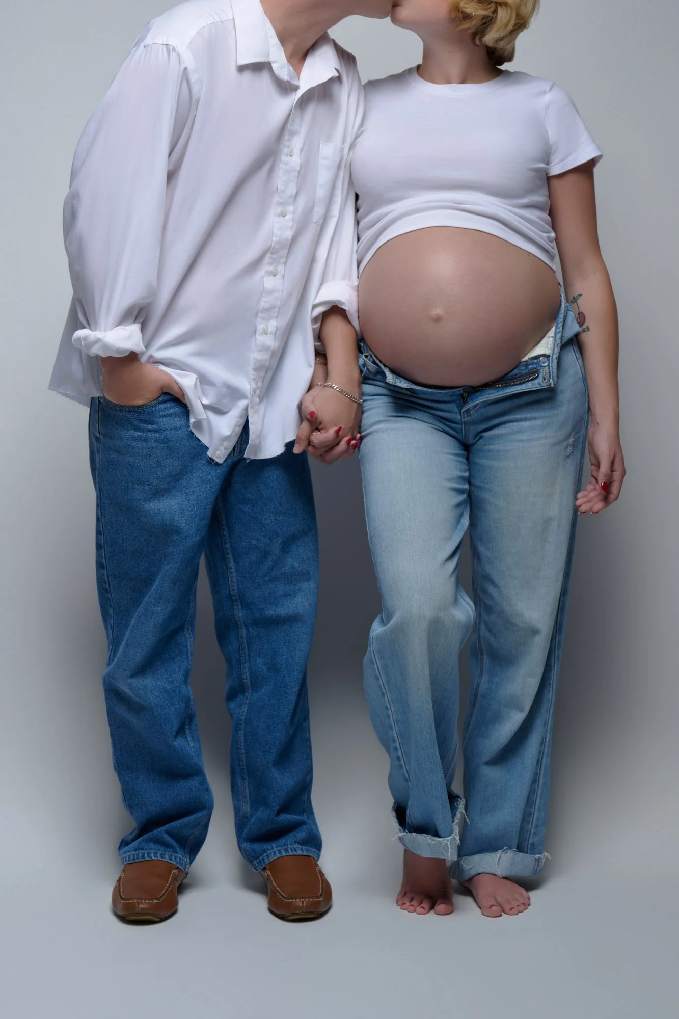 Kissing her partner in a crop tee and light jeans, holding hands beside the bump.