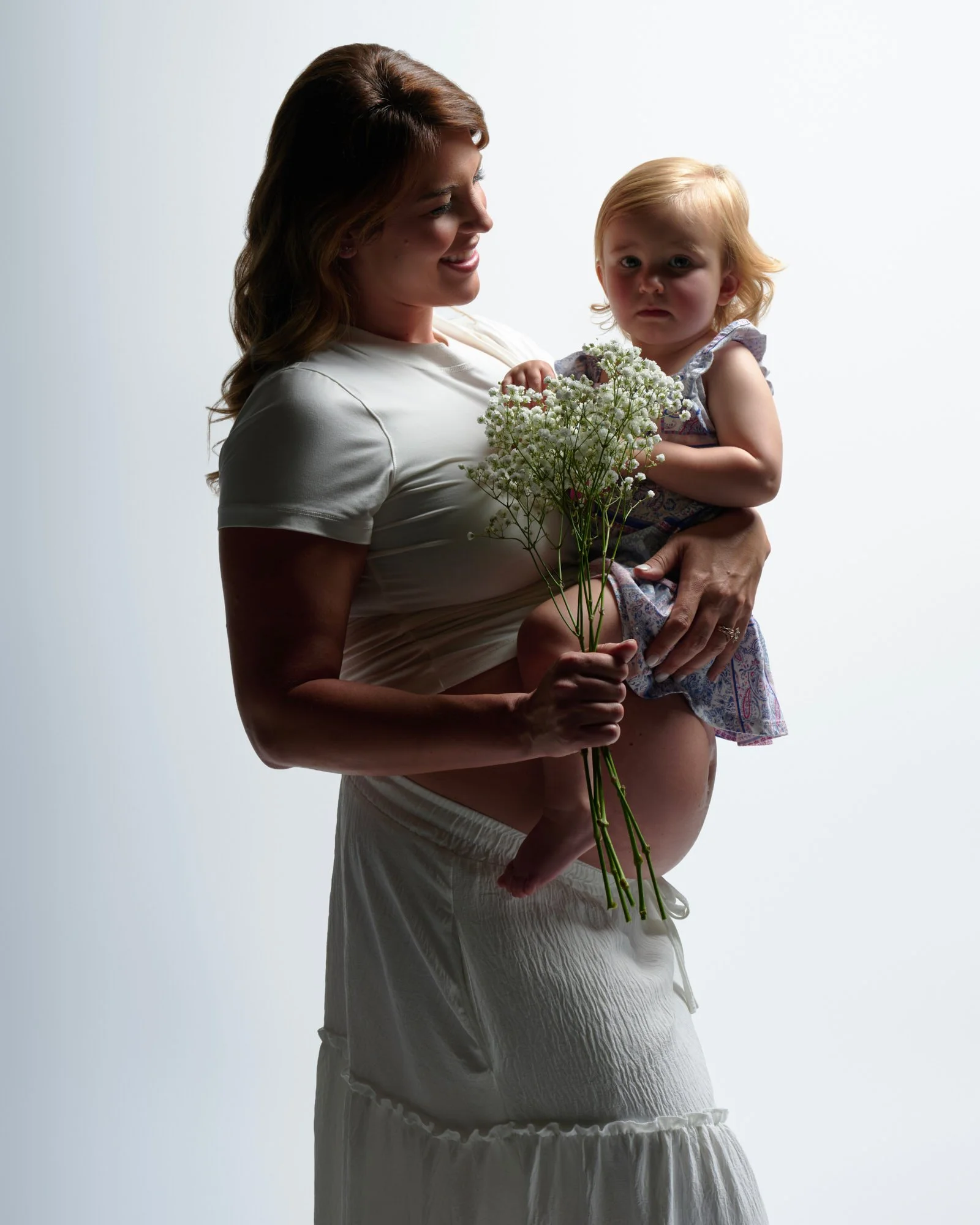 Mom holds her toddler on her hip with baby's breath flowers, backlit in white.