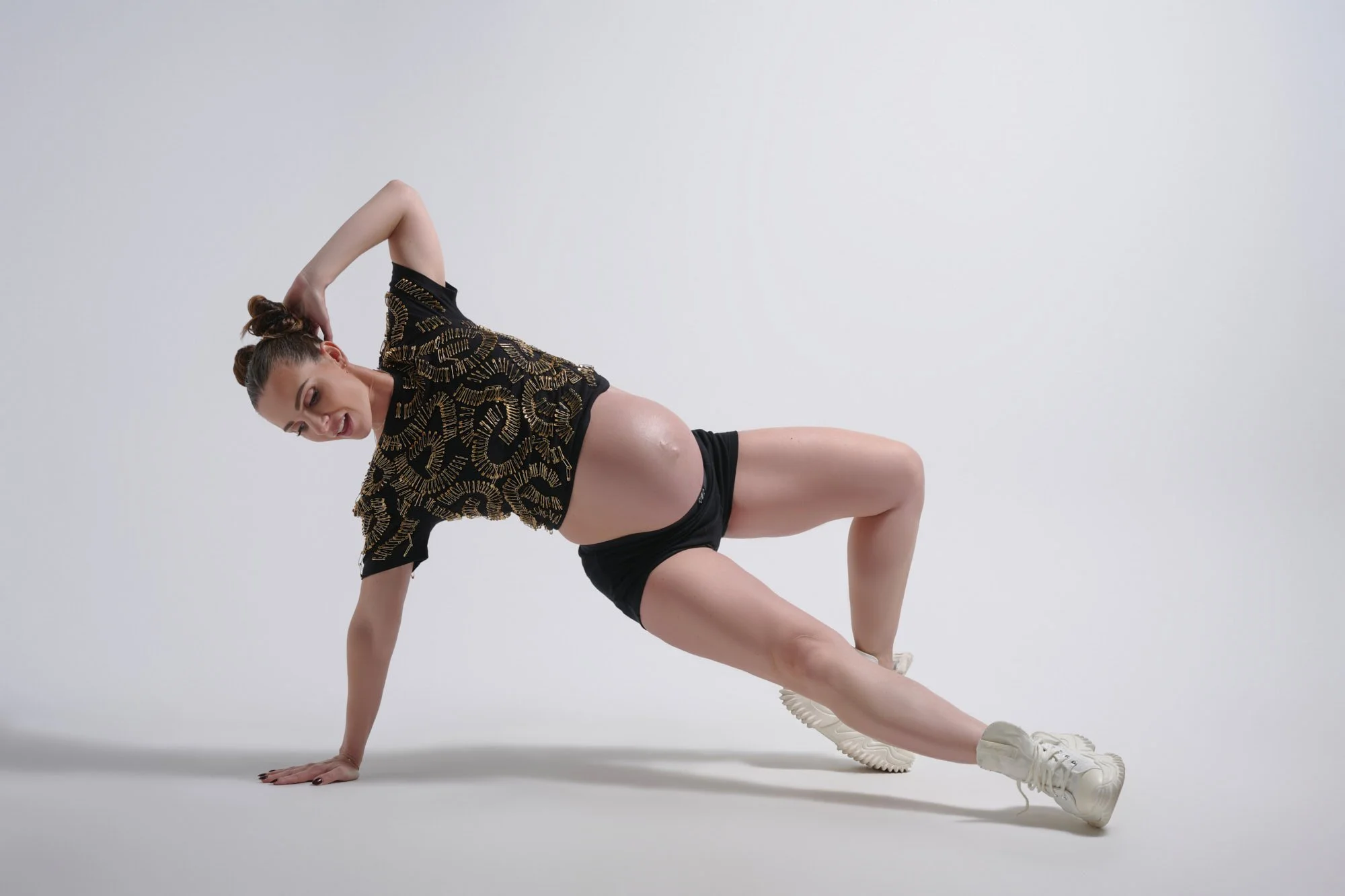 Side plank on a white studio floor in a gold-embroidered crop and sneakers, bump in profile.